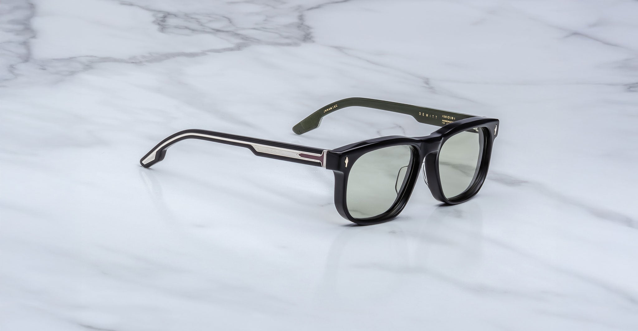 The Jacques Marie Mage Dewitt sunglasses feature a black rectangular frame with green-tinted lenses, black temples accented with metallic details, and olive green inner sides, resting stylishly on a white marble surface.