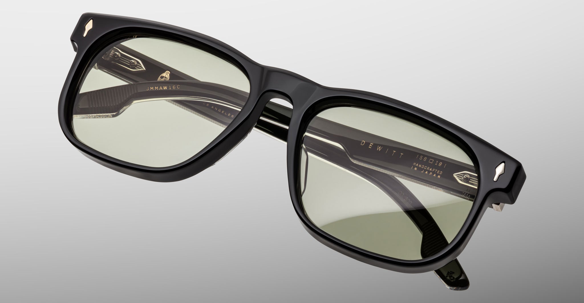 The Jacques Marie Mage Dewitt eyeglasses feature a black rectangular frame with light green tinted lenses, shown on a plain light gray background. The inside of the arms displays engraved text and small gold-colored accents.