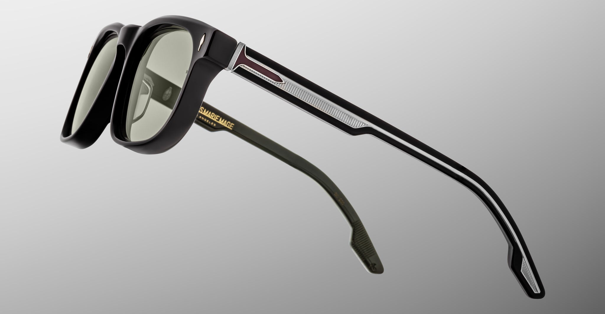 A close-up of Jacques Marie Mage Dewitt sunglasses in black with dark lenses, featuring textured silver accents and geometric temple patterns, set against a light gray gradient background.