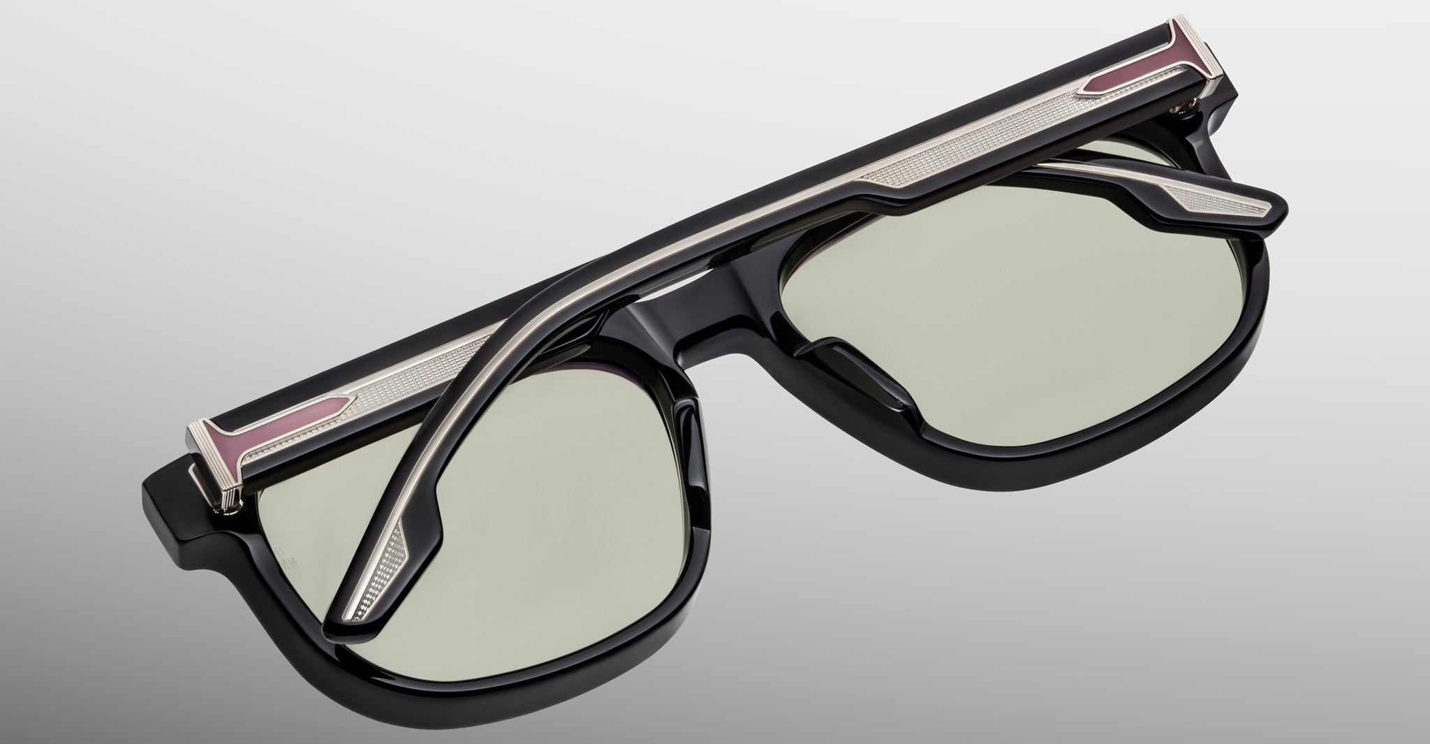 A close-up of Jacques Marie Mage Dewitt sunglasses featuring thick black frames, green-tinted lenses, and metallic accents on the temples and brow bar, set against a light gradient background.