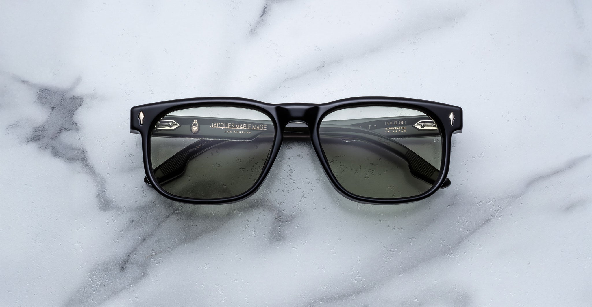A pair of Jacques Marie Mage Dewitt black rectangular sunglasses with dark lenses lies face up on a white marble surface with gray veining.