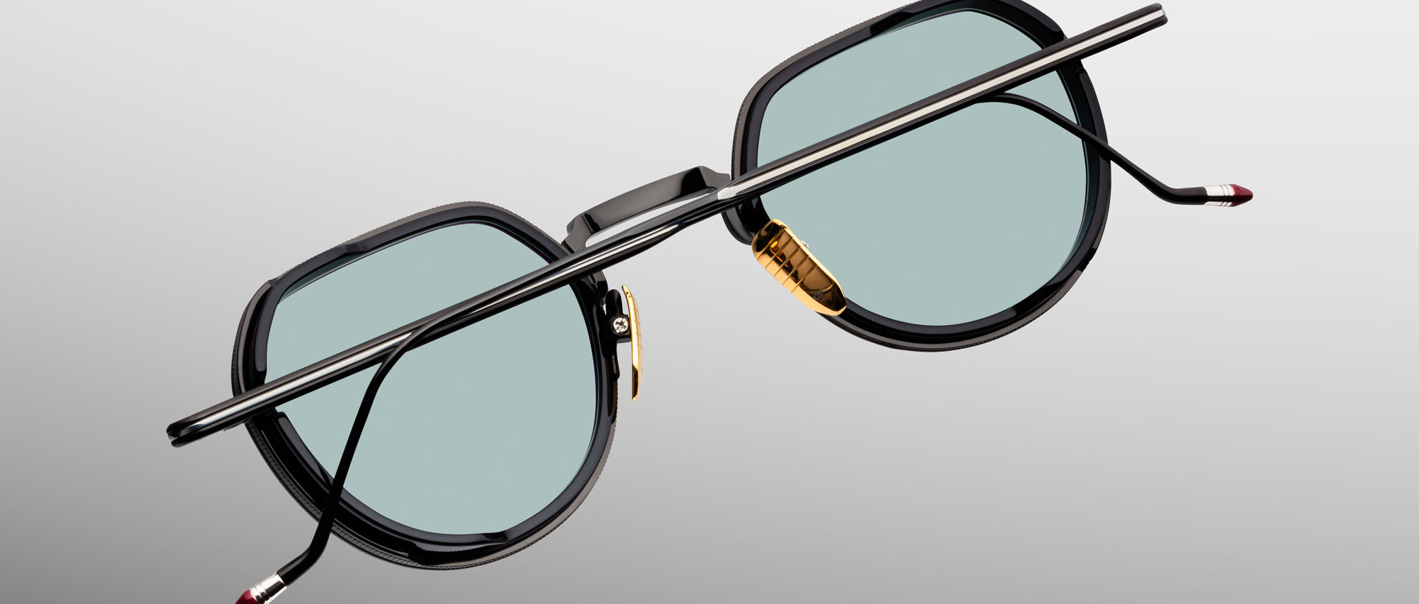 Jacques Marie Mage Dixon sunglasses with black frames, teal lenses, gold nose pads, and thin temples shown from the front.