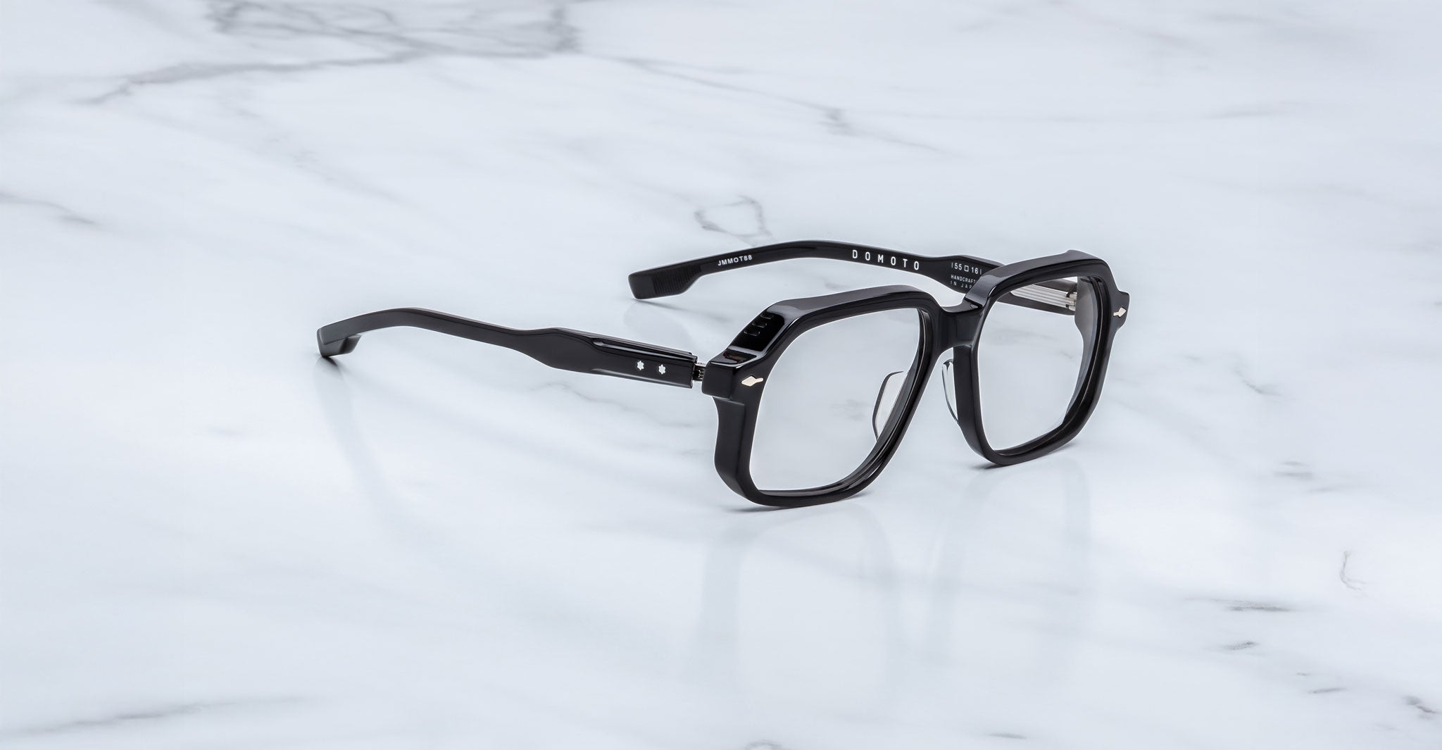 A pair of Jacques Marie Mage Domoto black, thick-rimmed rectangular eyeglasses with clear lenses and silver accents, handcrafted in Japan, rests on a white marble surface.