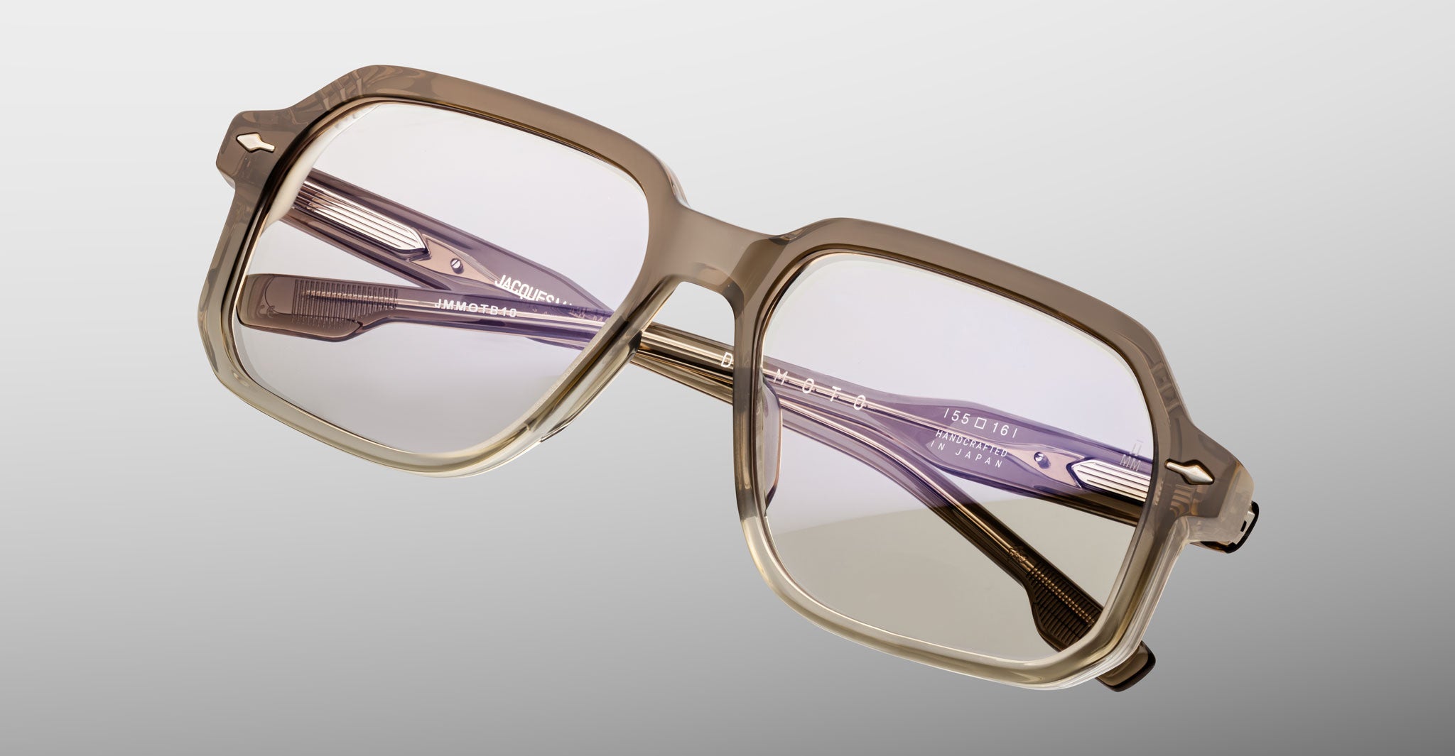 A pair of Jacques Marie Mage Domoto eyeglasses, handcrafted in Japan with square transparent taupe frames and light-tinted lenses, displayed at an angle on a light gray background.
