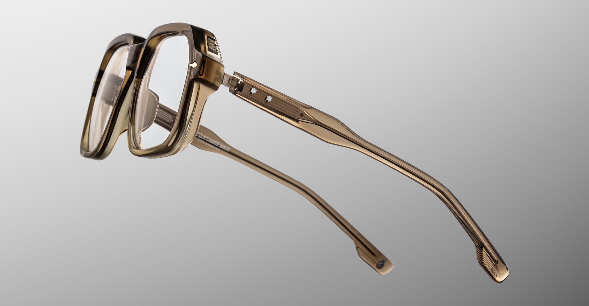 The Jacques Marie Mage Domoto eyeglasses feature stylish, thick transparent brown frames with wide temples and metal accents, handcrafted in Japan and displayed on a plain gray gradient background.