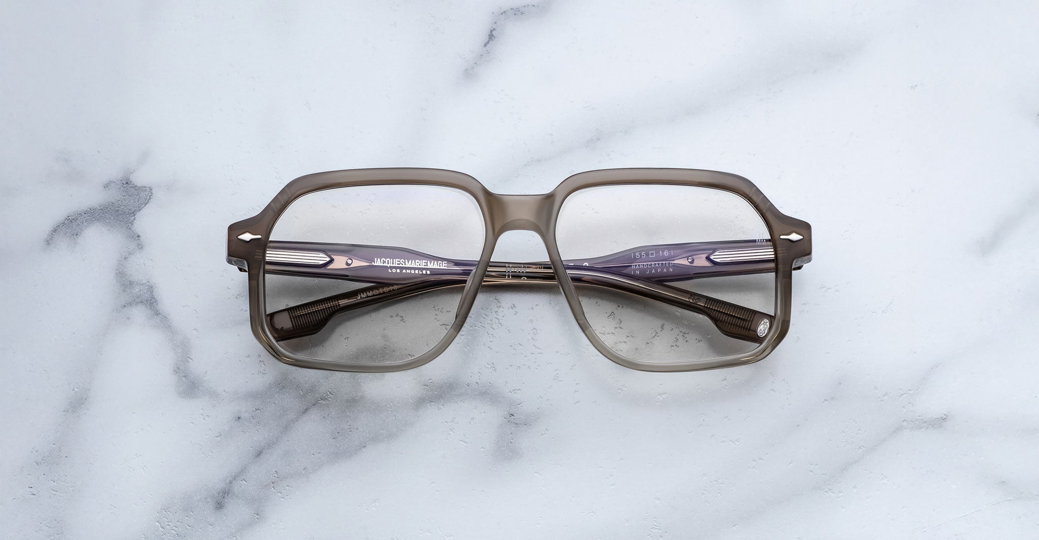 The Jacques Marie Mage Domoto is a pair of large, square eyeglasses handcrafted in Japan with a light brown translucent frame and clear lenses, shown face-down on white marble with subtle gray veining.