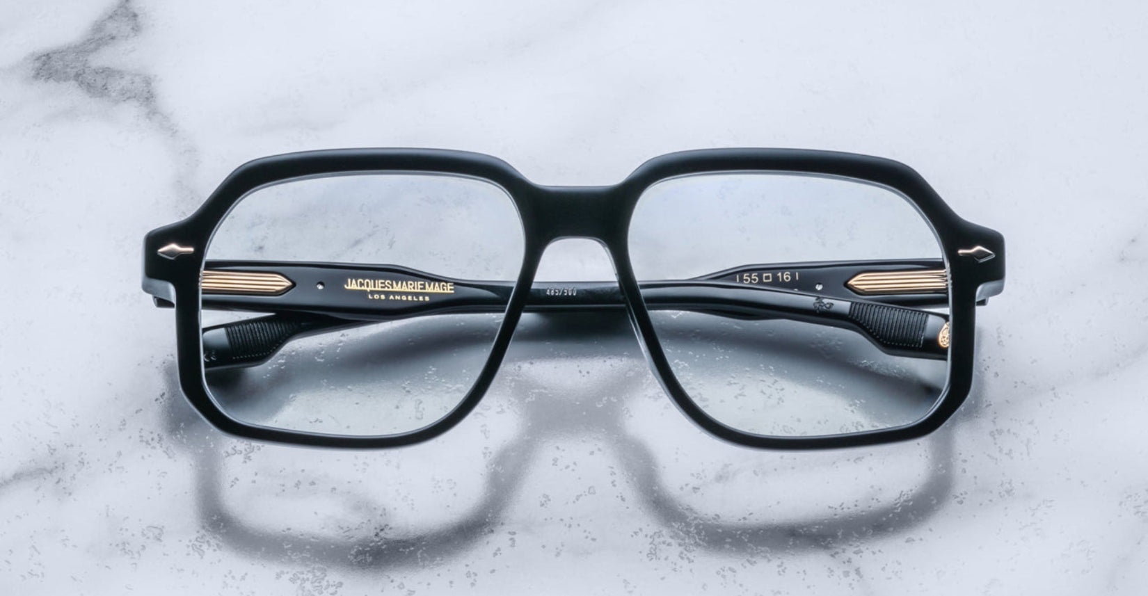 A pair of Jacques Marie Mage Domoto black rectangular eyeglasses with gold accents rests on a white marble surface, viewed from above with the arms crossed behind the lenses.