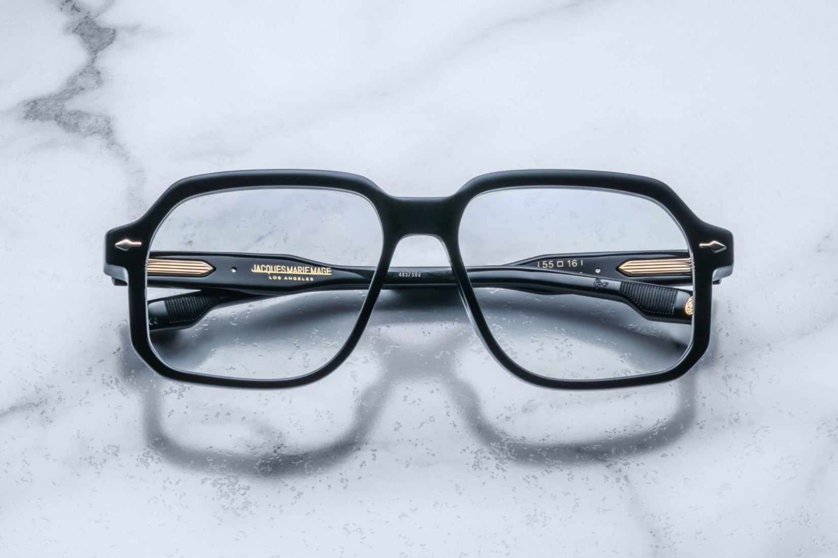 A pair of Jacques Marie Mage Domoto black rectangular eyeglasses with gold accents rests on a white marble surface, viewed from above with the arms crossed behind the lenses.