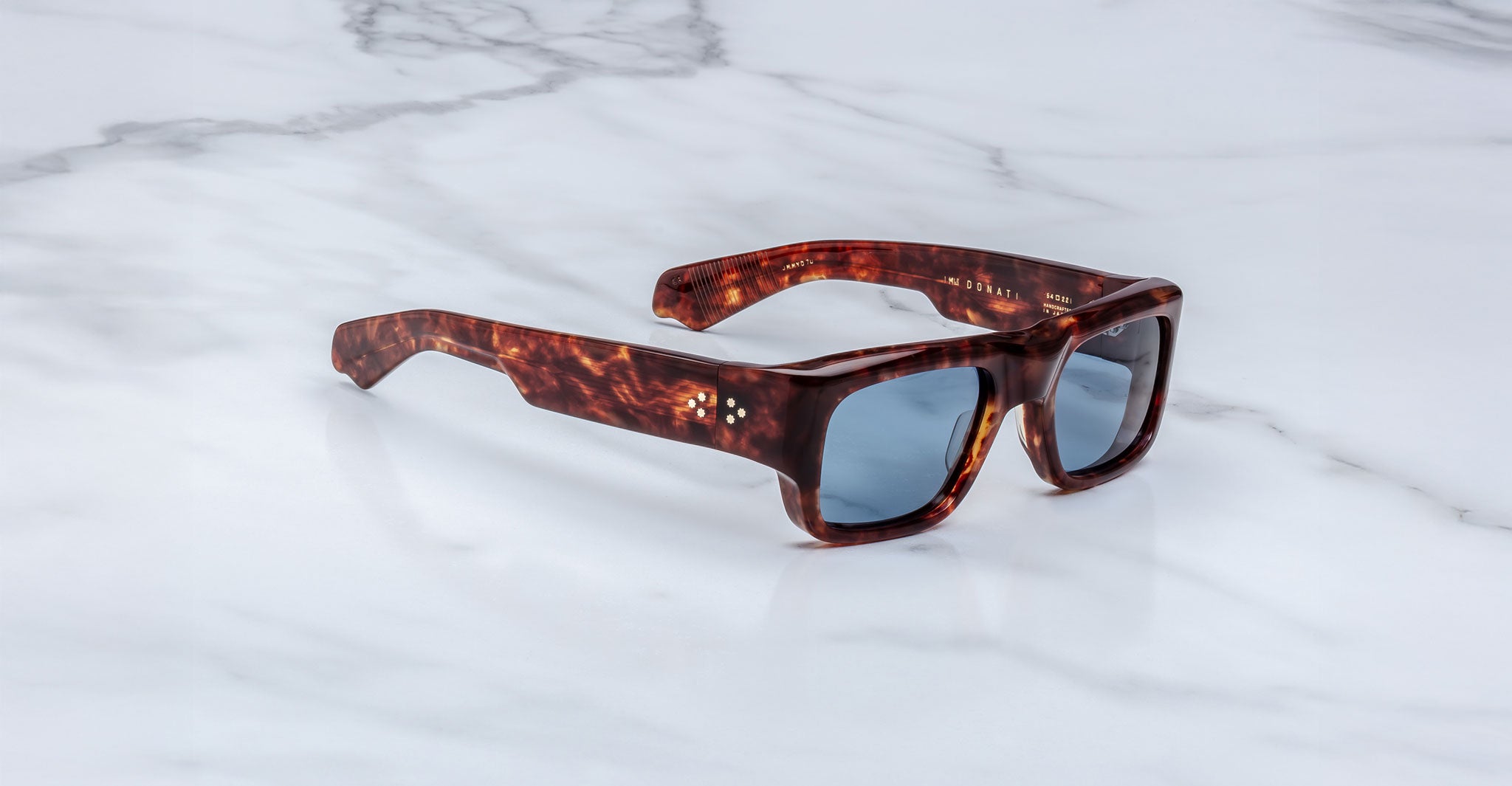 The Jacques Marie Mage Donat sunglasses, featuring thick tortoiseshell rectangular frames and dark lenses, rest on a white marble surface.