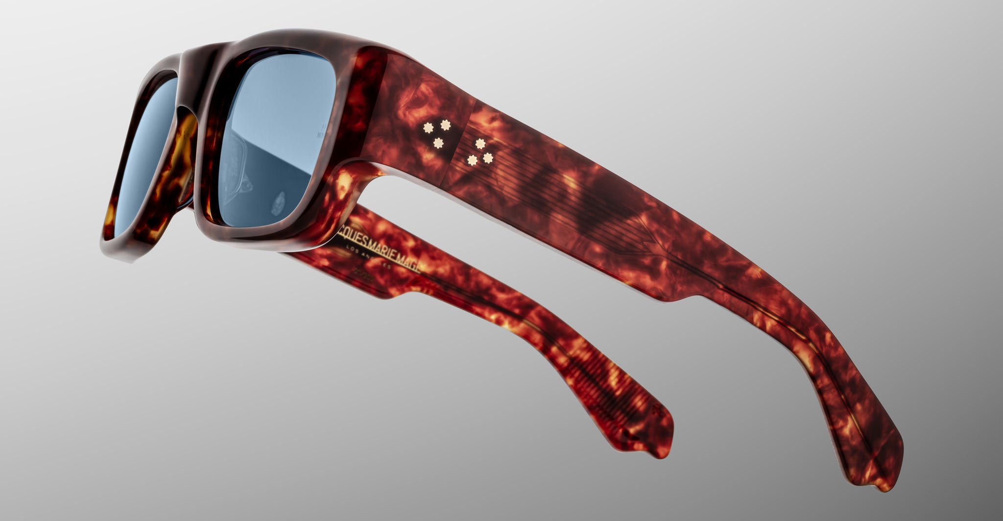 The Jacques Marie Mage Donat sunglasses feature thick, glossy brown tortoiseshell frames with blue-tinted lenses, four white dots on the temple, and gold text on the arm, shown against a light gray background.