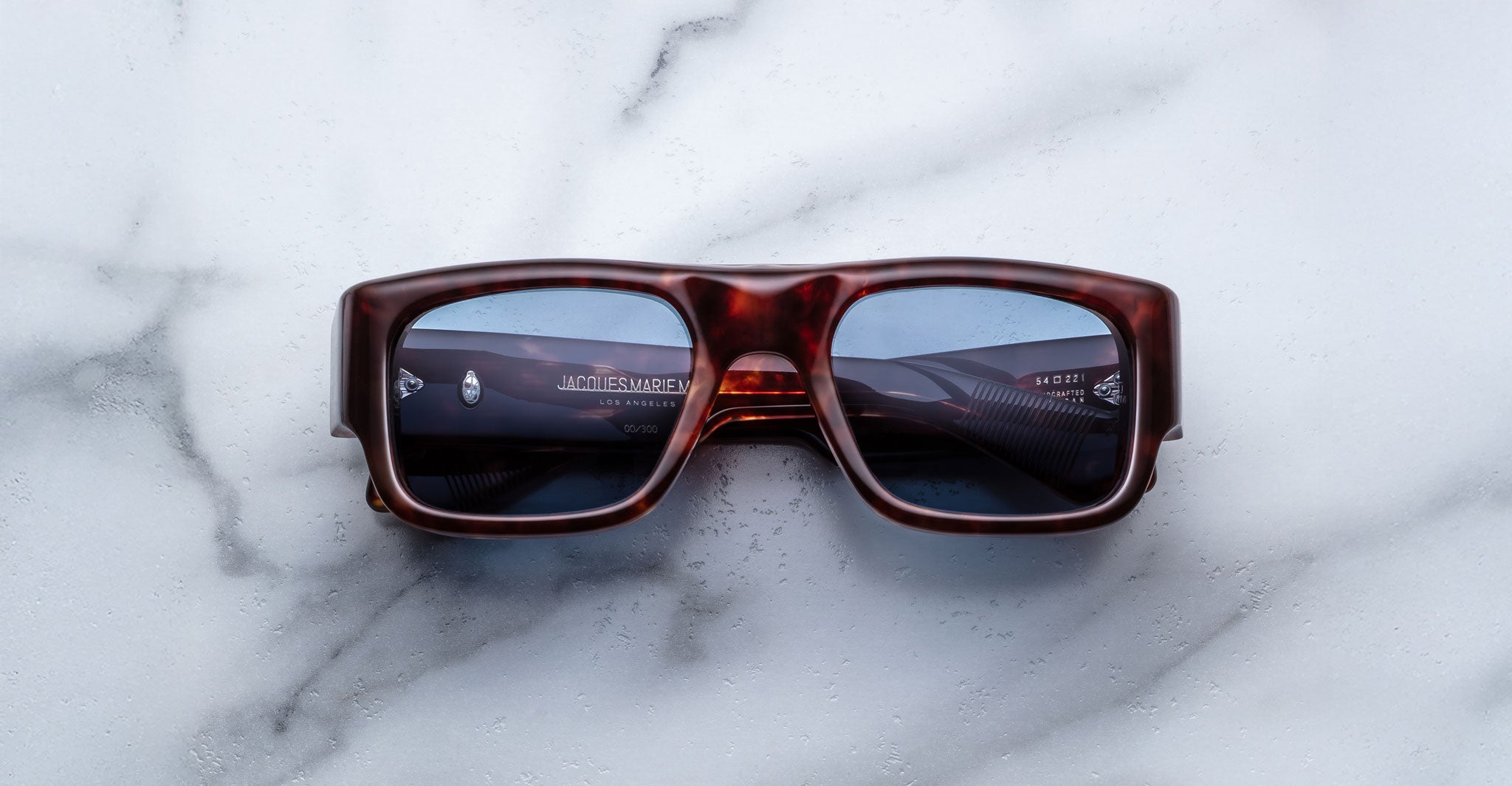 The Jacques Marie Mage Donat rectangular sunglasses, featuring a thick dark tortoiseshell frame and blue-tinted lenses, rest on a white marble surface.