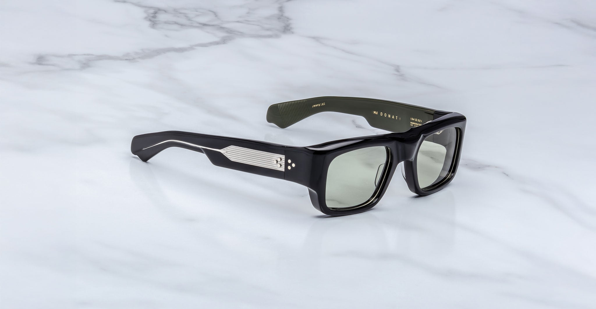 The Jacques Marie Mage Donat sunglasses feature black rectangular frames, tinted lenses, metallic arm detailing, and dark green interior arms, and are displayed on a white marble surface.
