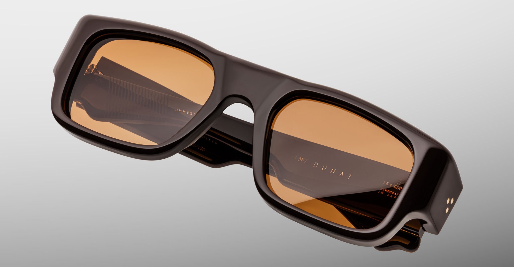 The Jacques Marie Mage Donat sunglasses feature a rectangular shape, thick black frames, and amber-tinted lenses, shown against a light gray background.