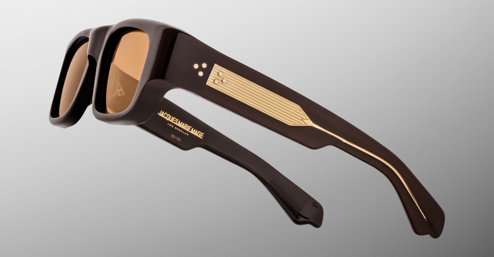 A close-up of Jacques Marie Mage Donat sunglasses with thick black frames, gold detailing on the temples, and decorative gold lines and text reading ACKERMAN & JANCE, set against a neutral gray background.