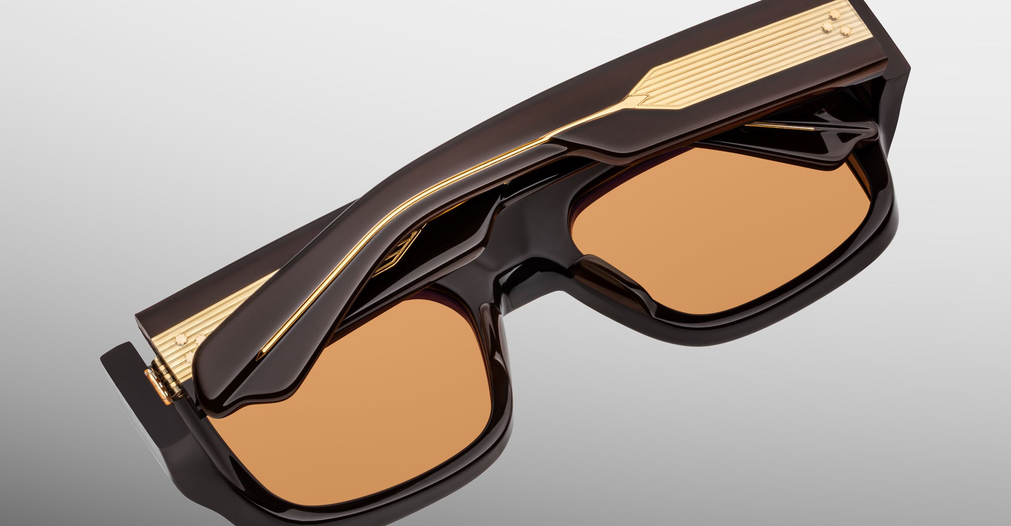 The Jacques Marie Mage Donat sunglasses feature rectangular frames in glossy black acetate with gold detailing on the arms and top, resting on a white surface. The amber-tinted lenses complete their sophisticated look.