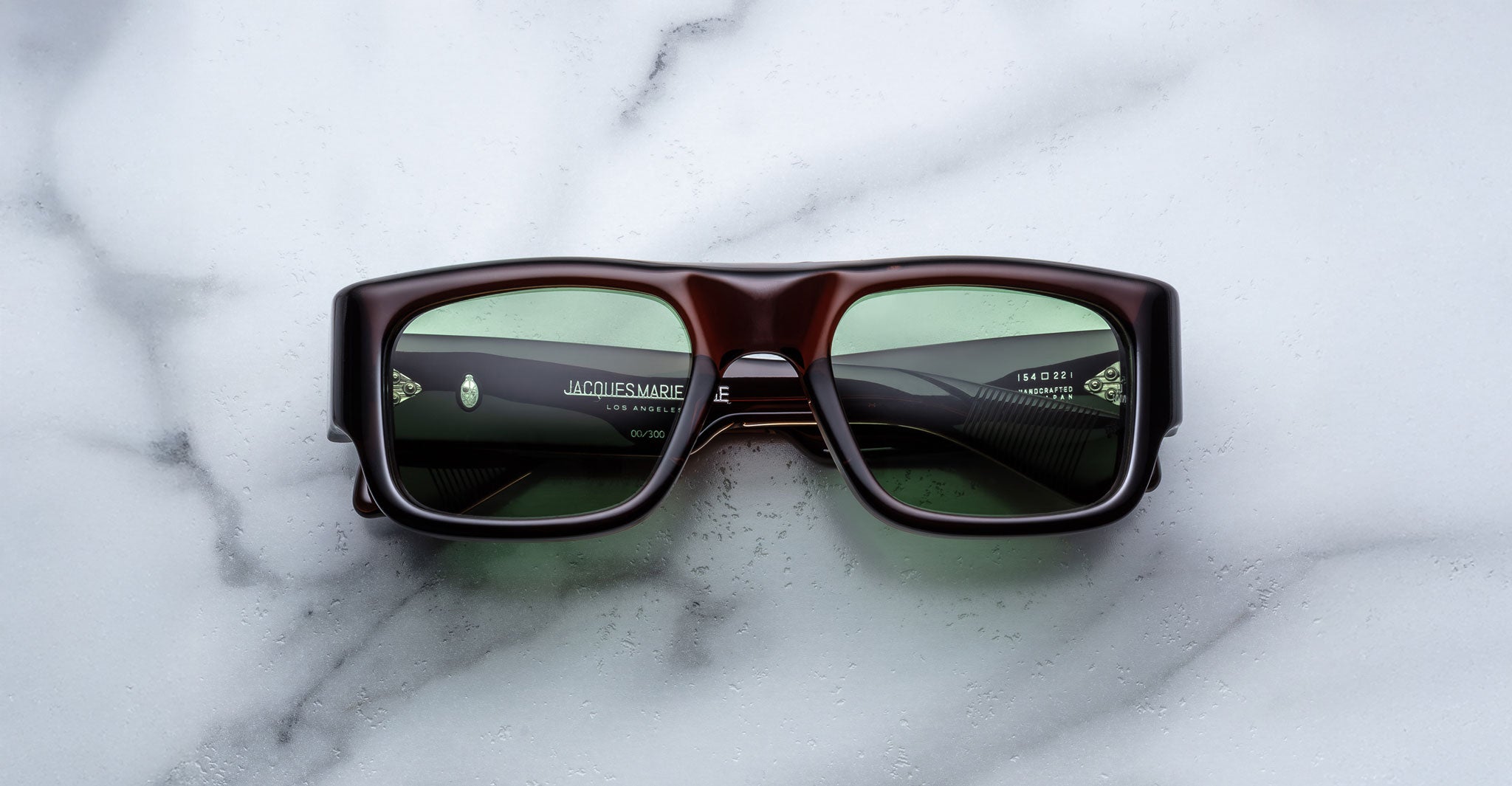 A pair of Jacques Marie Mage Donat rectangular, chunky dark brown sunglasses with green-tinted lenses rests on a white marble surface, with brand and model details visible inside the left arm.