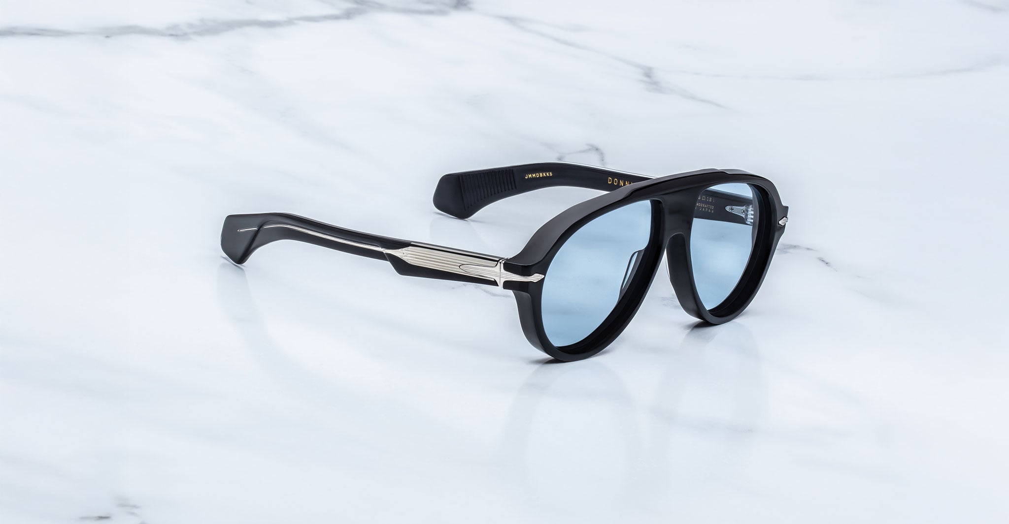 Jacques Marie Mage Donnybrooke sunglasses with blue-tinted lenses and silver arm detailing, limited-edition frames by Jacques Marie Mage, rest on a white marble surface.
