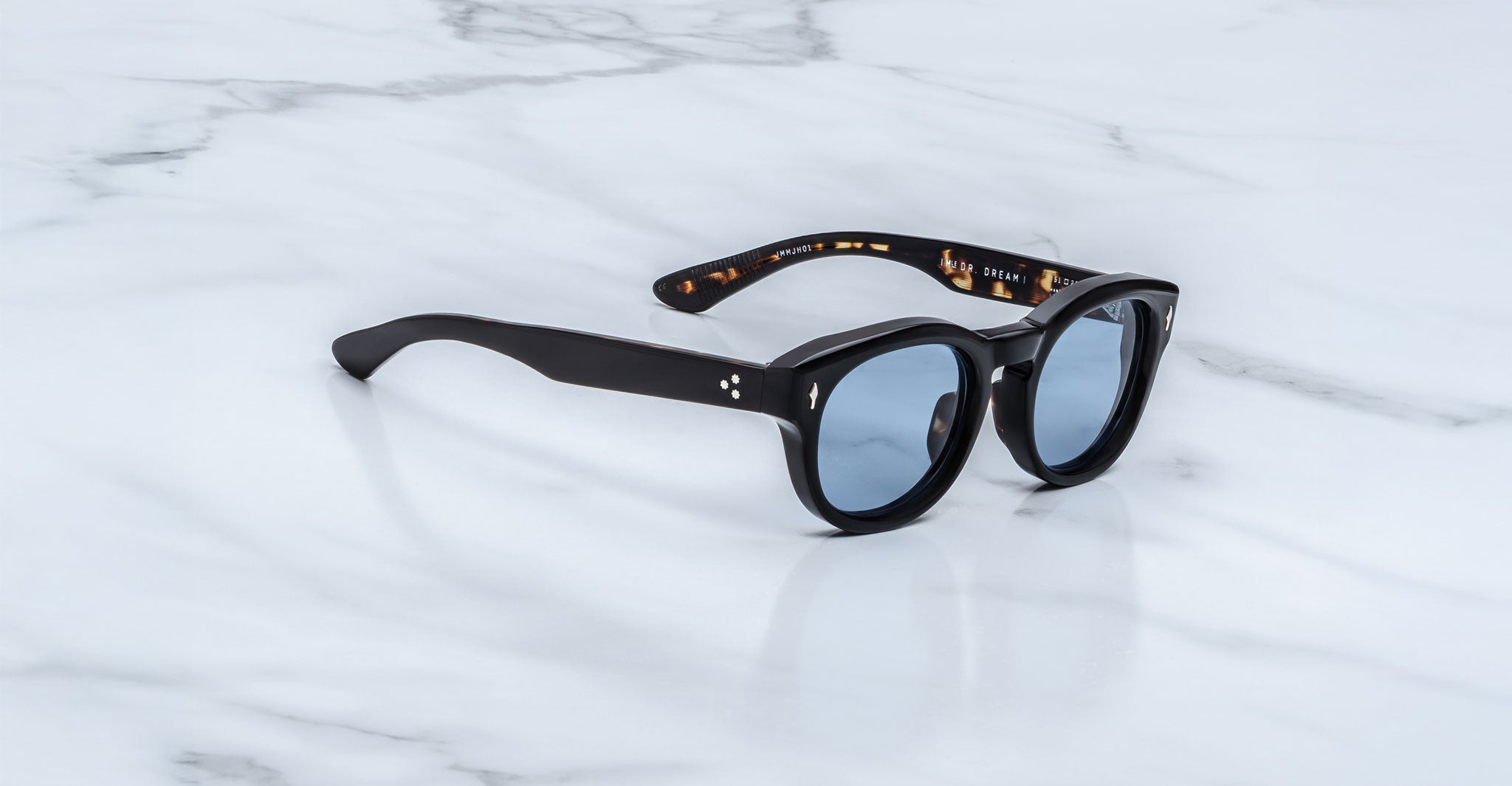 Jacques Marie Mage Dr. Dream sunglasses—black, round frames with blue lenses—are folded on a white marble surface, highlighting the luxury and Japanese craftsmanship of Jacques Marie Mage eyewear.