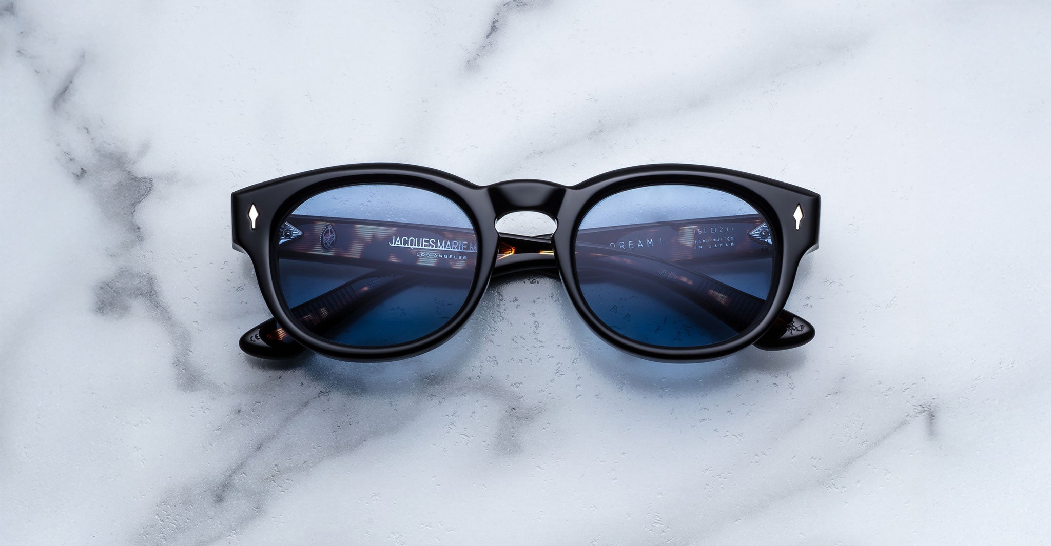 The Jacques Marie Mage Dr. Dream sunglasses feature black round frames and blue tinted lenses, displayed on white marble with the lenses reflecting a computer screen—an elegant, luxurious eyewear choice.