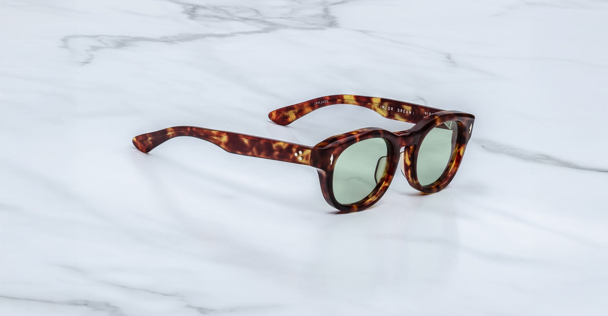 The Jacques Marie Mage Dr. Dream sunglasses by Jacques Marie Mage, featuring a tortoiseshell pattern and round green-tinted lenses, rest on a white marble surface.