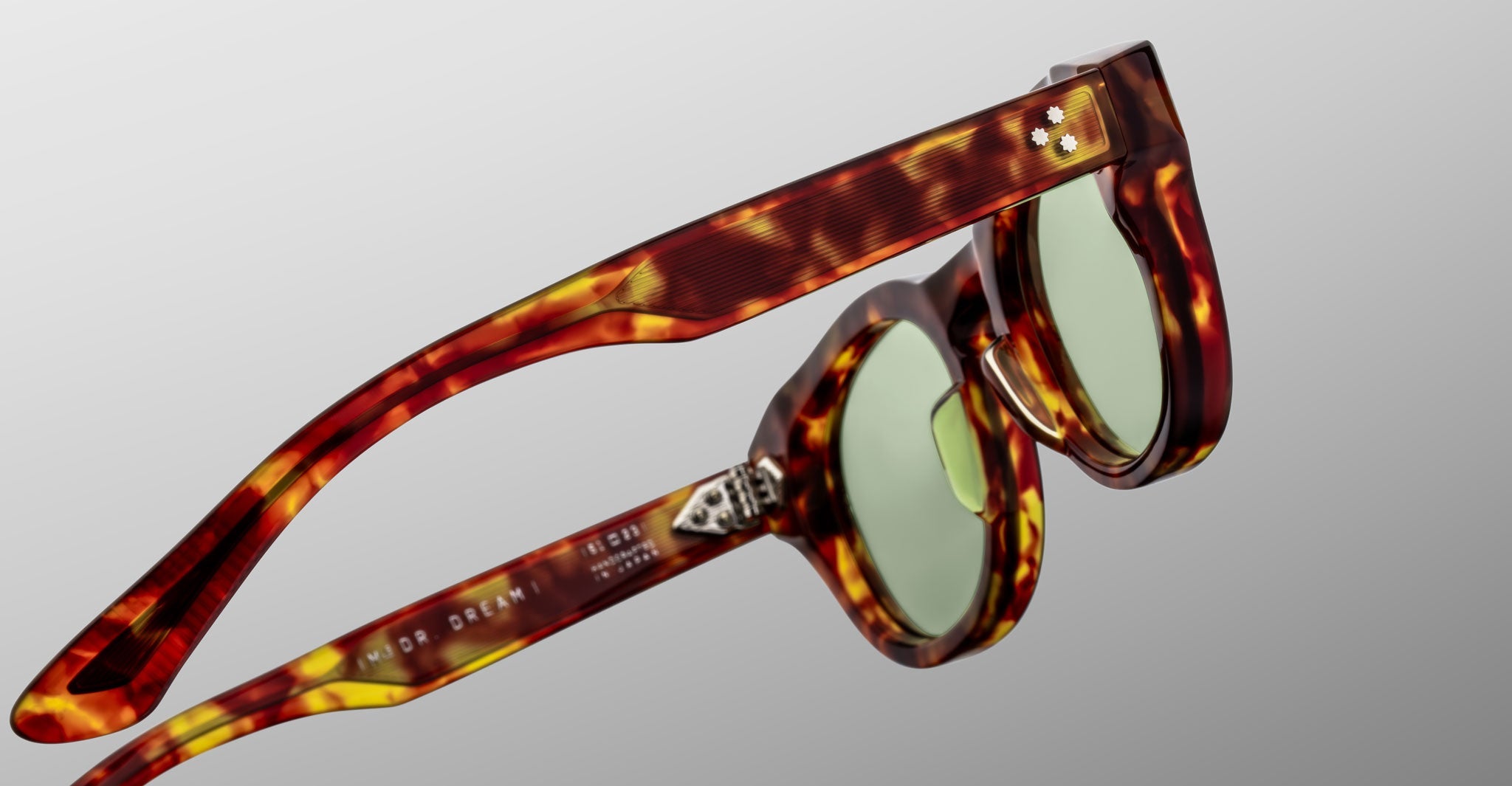 Close-up of Jacques Marie Mage Dr. Dream sunglasses by Jacques Marie Mage, featuring tortoiseshell frames and green lenses, positioned to highlight the side and front against a gray background.