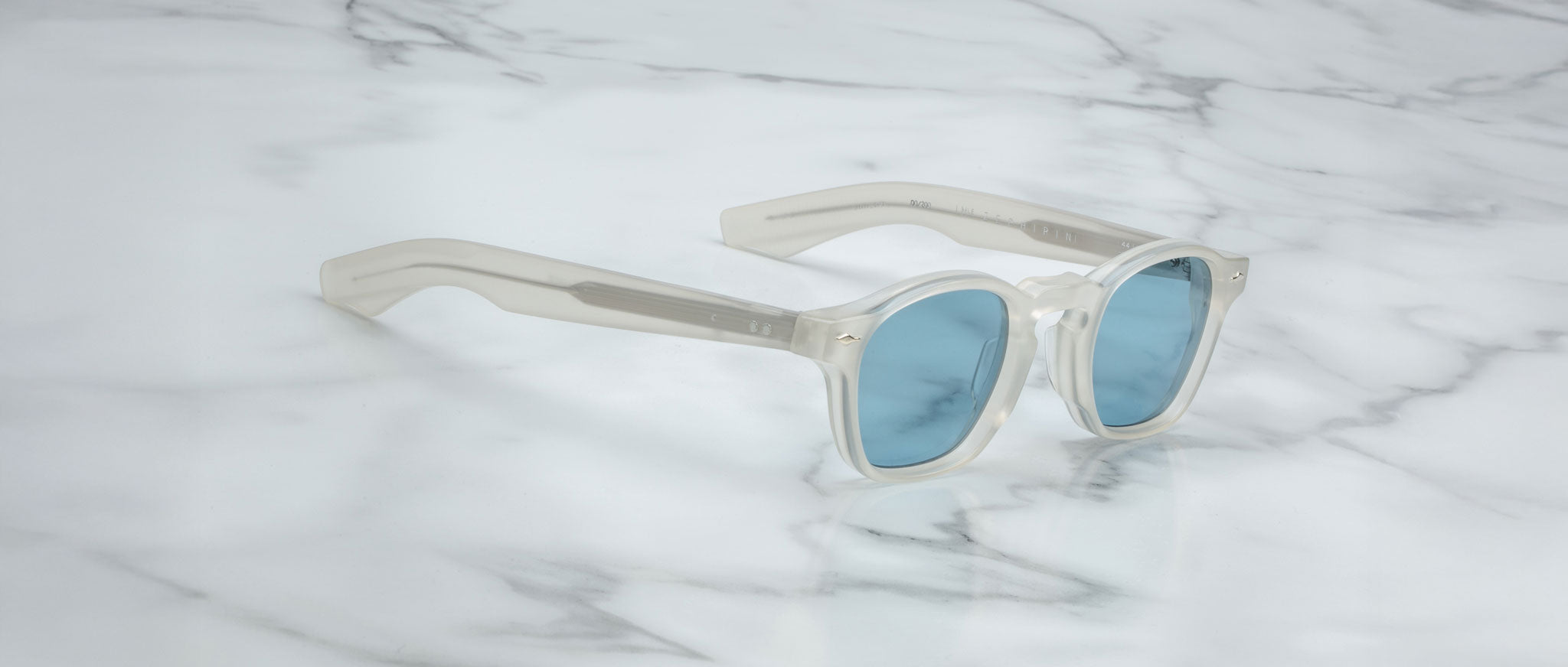The Jacques Marie Mage Zephirin sunglasses by Jacques Marie Mage, featuring translucent white frames and blue lenses, inspired by handcrafted acetate designs, are displayed elegantly on a white marble surface.