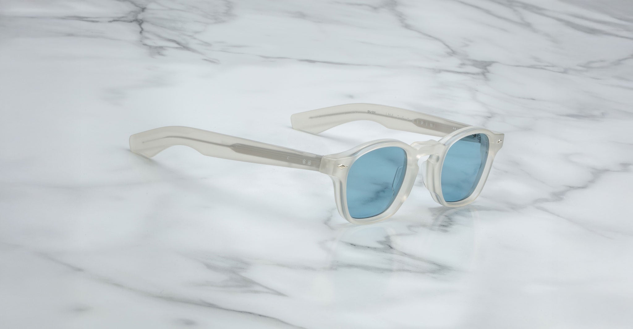 The Jacques Marie Mage Zephirin sunglasses by Jacques Marie Mage, featuring translucent white frames and blue lenses, inspired by handcrafted acetate designs, are displayed elegantly on a white marble surface.