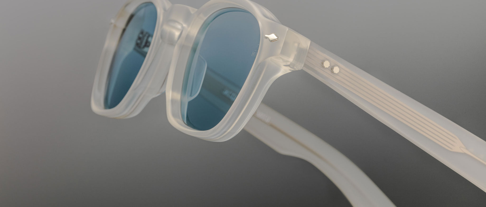 Close-up of Jacques Marie Mage Zephirin limited edition eyewear, showcasing translucent handcrafted acetate frames with blue-tinted lenses and metallic details, by Jacques Marie Mage, set against a neutral background.