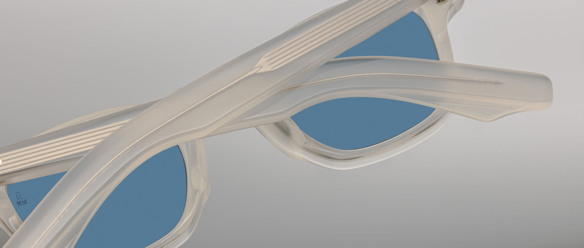 Close-up of Jacques Marie Mage Zephirin sunglasses in translucent white with blue lenses, shown angled with crossed arms—a stunning, limited edition model from Jacques Marie Mage.