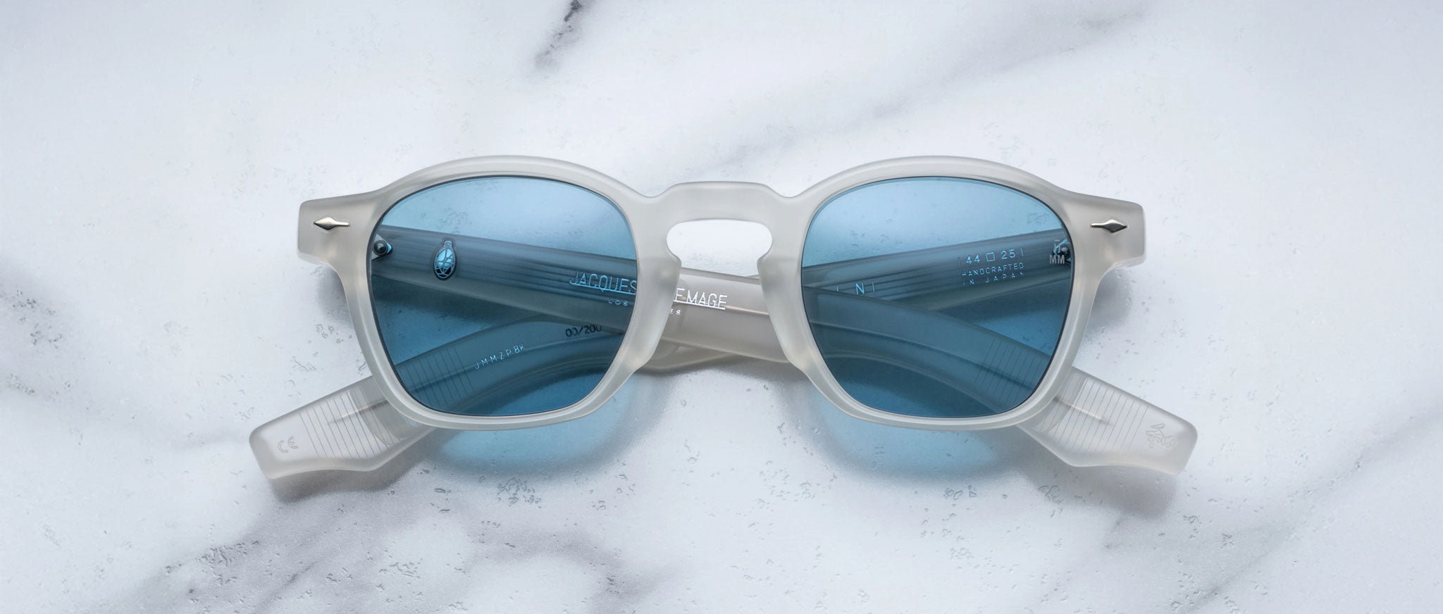 Limited edition Jacques Marie Mage Zephirin sunglasses feature a translucent white acetate frame and blue-tinted lenses, capturing the elegance of handcrafted eyewear. Displayed on a white marble surface by Jacques Marie Mage.
