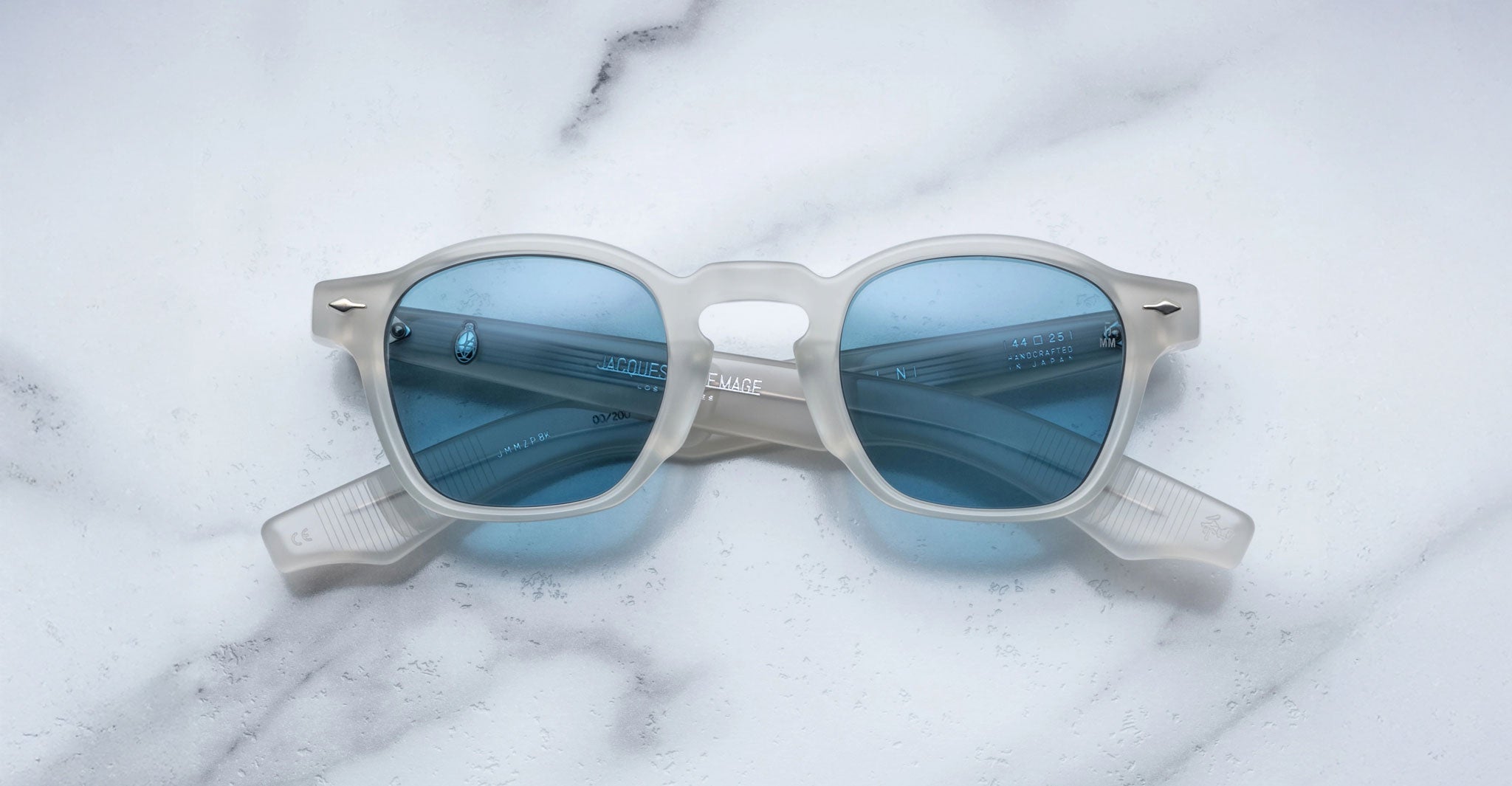 Limited edition Jacques Marie Mage Zephirin sunglasses feature a translucent white acetate frame and blue-tinted lenses, capturing the elegance of handcrafted eyewear. Displayed on a white marble surface by Jacques Marie Mage.