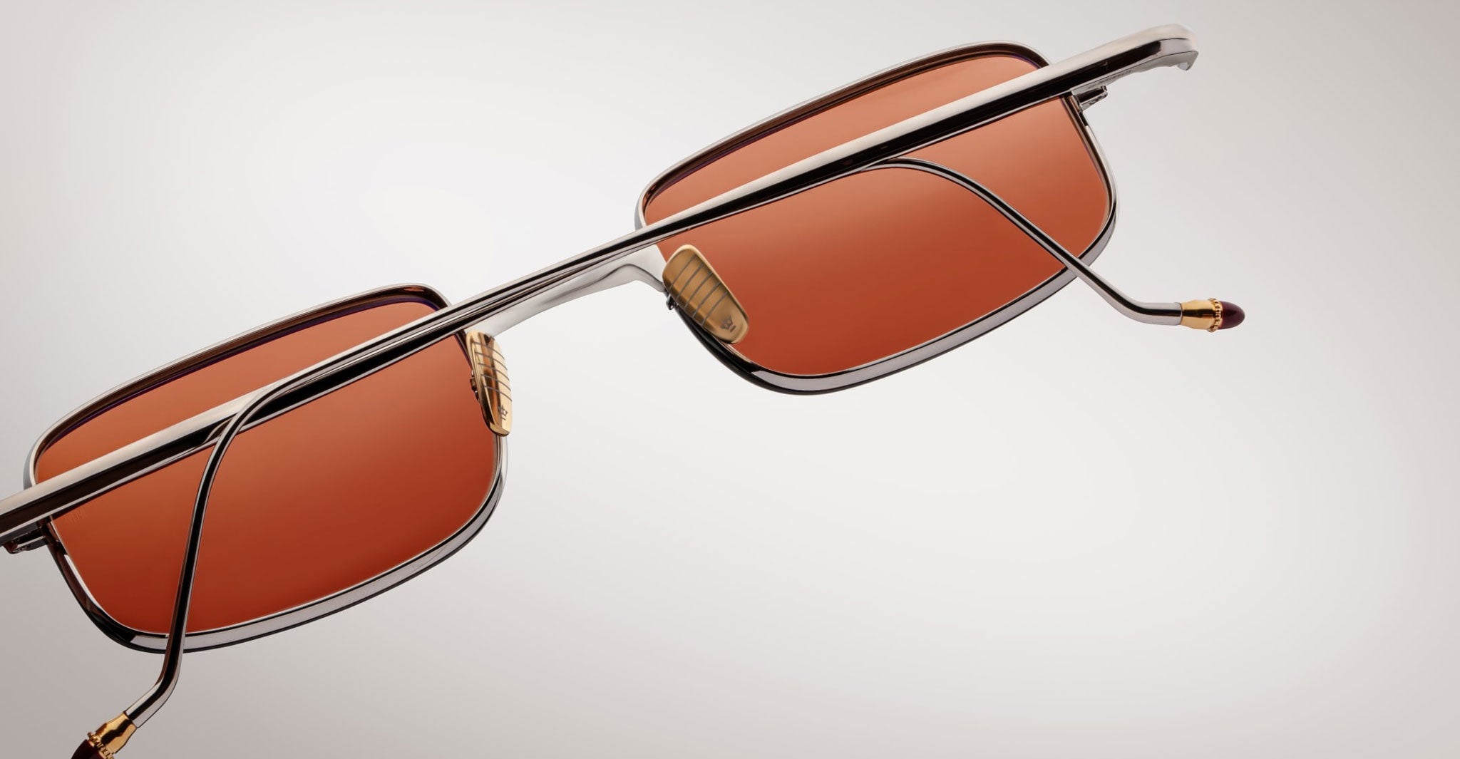 A close-up of Jacques Marie Mage Dusk sunglasses featuring rectangular reddish-brown lenses and a metallic frame, set against a light gray background.