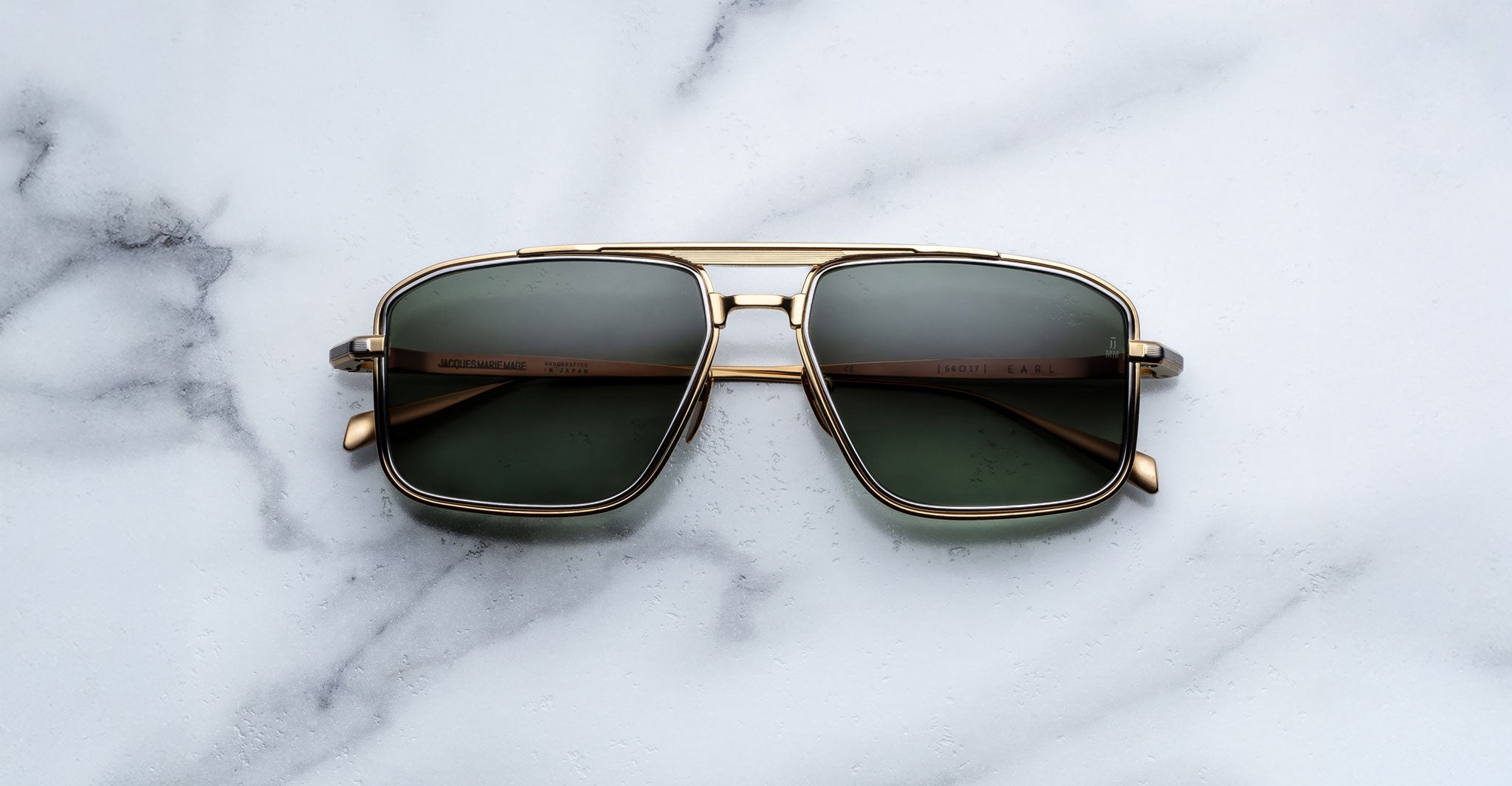 The Jacques Marie Mage Earl sunglasses, featuring gold frames, dark lenses, and Art Deco details, are displayed on white marble with grey veining.
