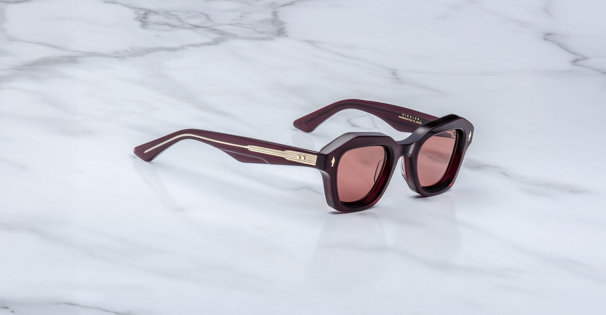 The Jacques Marie Mage Eichler is a pair of dark brown square sunglasses with gold accents on the arms, tinted brown lenses, and thick frames, shown resting on a white marble surface.