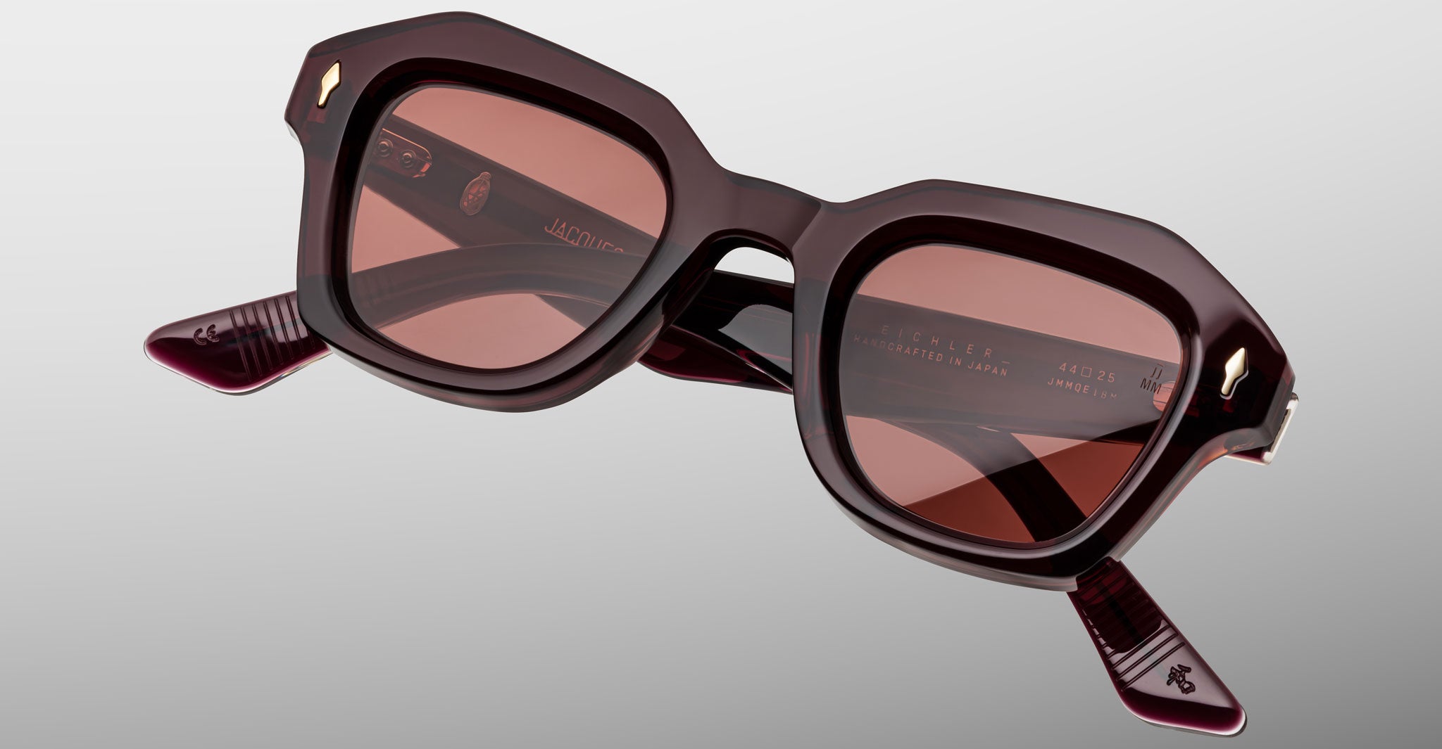 The Jacques Marie Mage Eichler sunglasses feature oversized square frames in dark burgundy with geometric edges, tinted lenses, subtle patterned arms, and gold-colored accents near the hinges.
