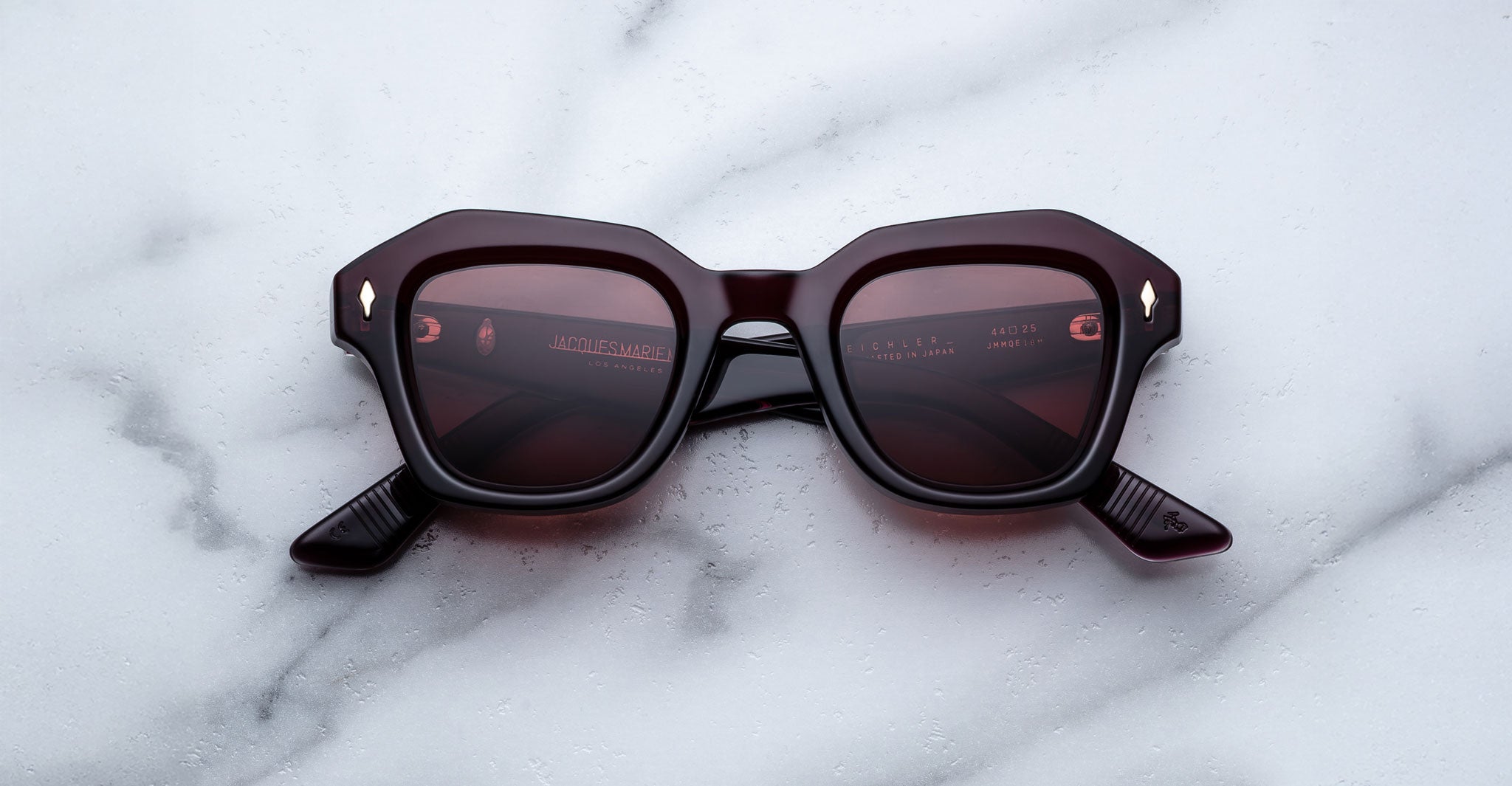 The Jacques Marie Mage Eichler sunglasses feature dark burgundy, thick geometric frames with tinted lenses and are displayed on a white marble surface.