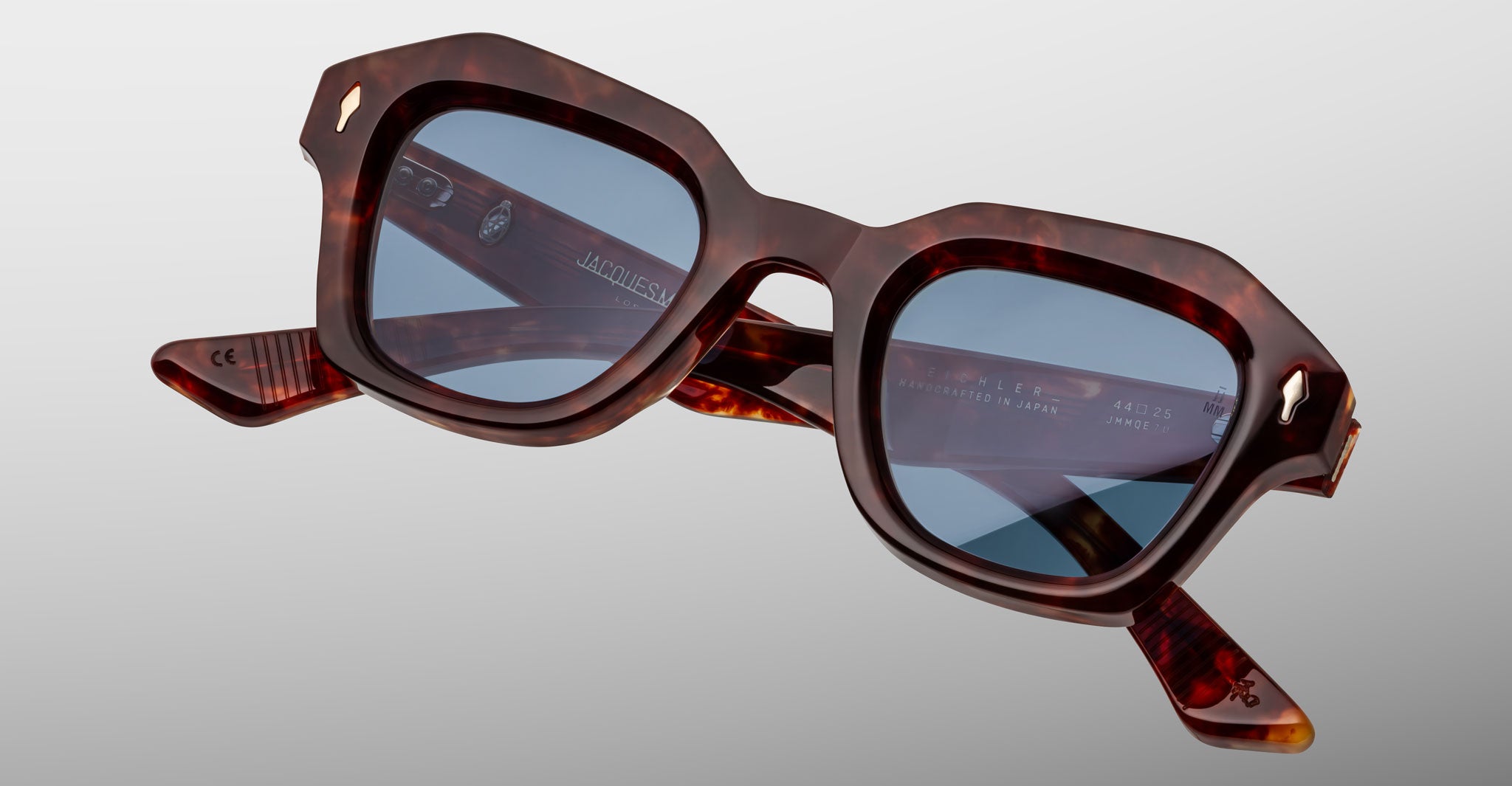 The Jacques Marie Mage Eichler sunglasses, featuring thick angular tortoiseshell frames and blue-tinted lenses, are displayed against a plain light gray background.