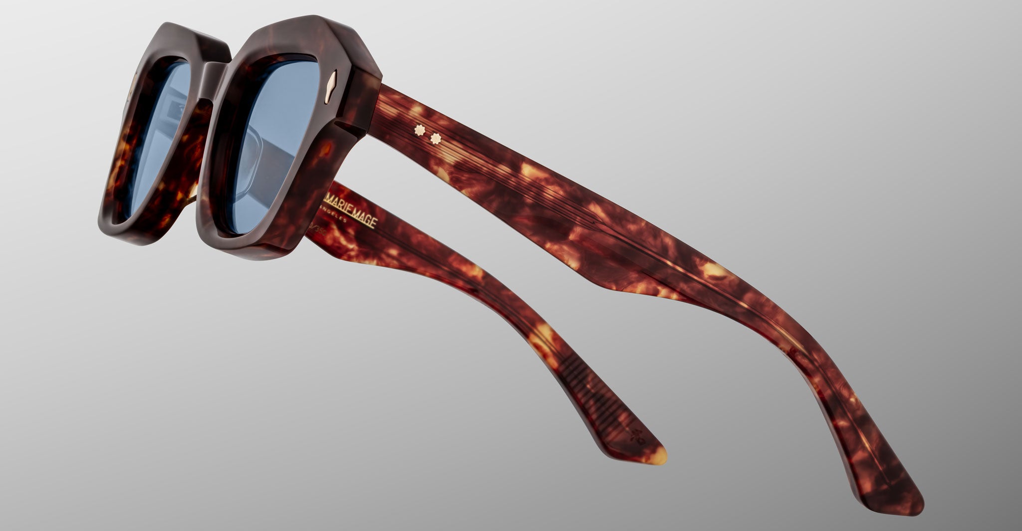 A close-up side view of the Jacques Marie Mage Eichler sunglasses, featuring dark tortoiseshell frames, blue-tinted lenses, and gold accents, displayed against a plain gradient background.
