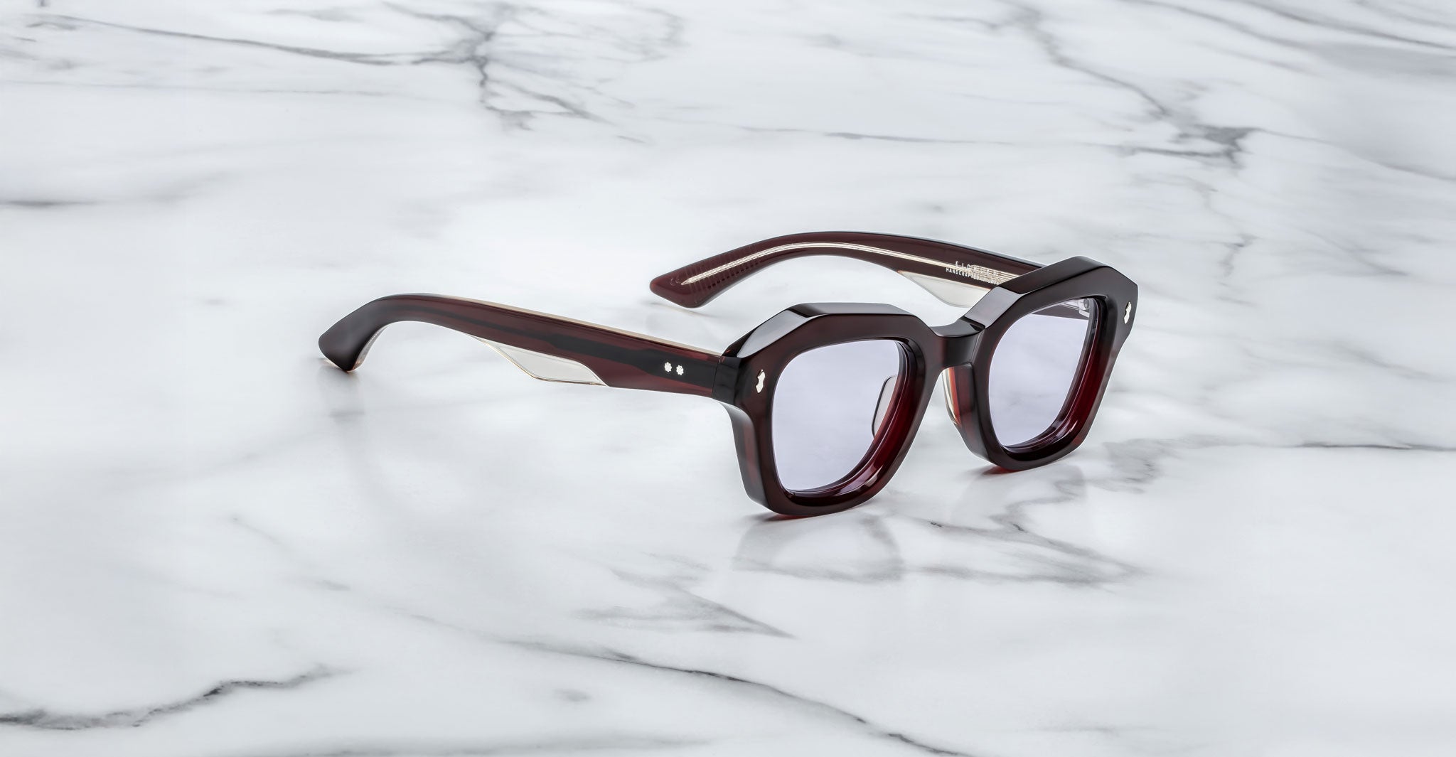 A pair of Jacques Marie Mage Eichler cherry color, thick-framed sunglasses with grey lenses sits on a white marble surface with lilac veins