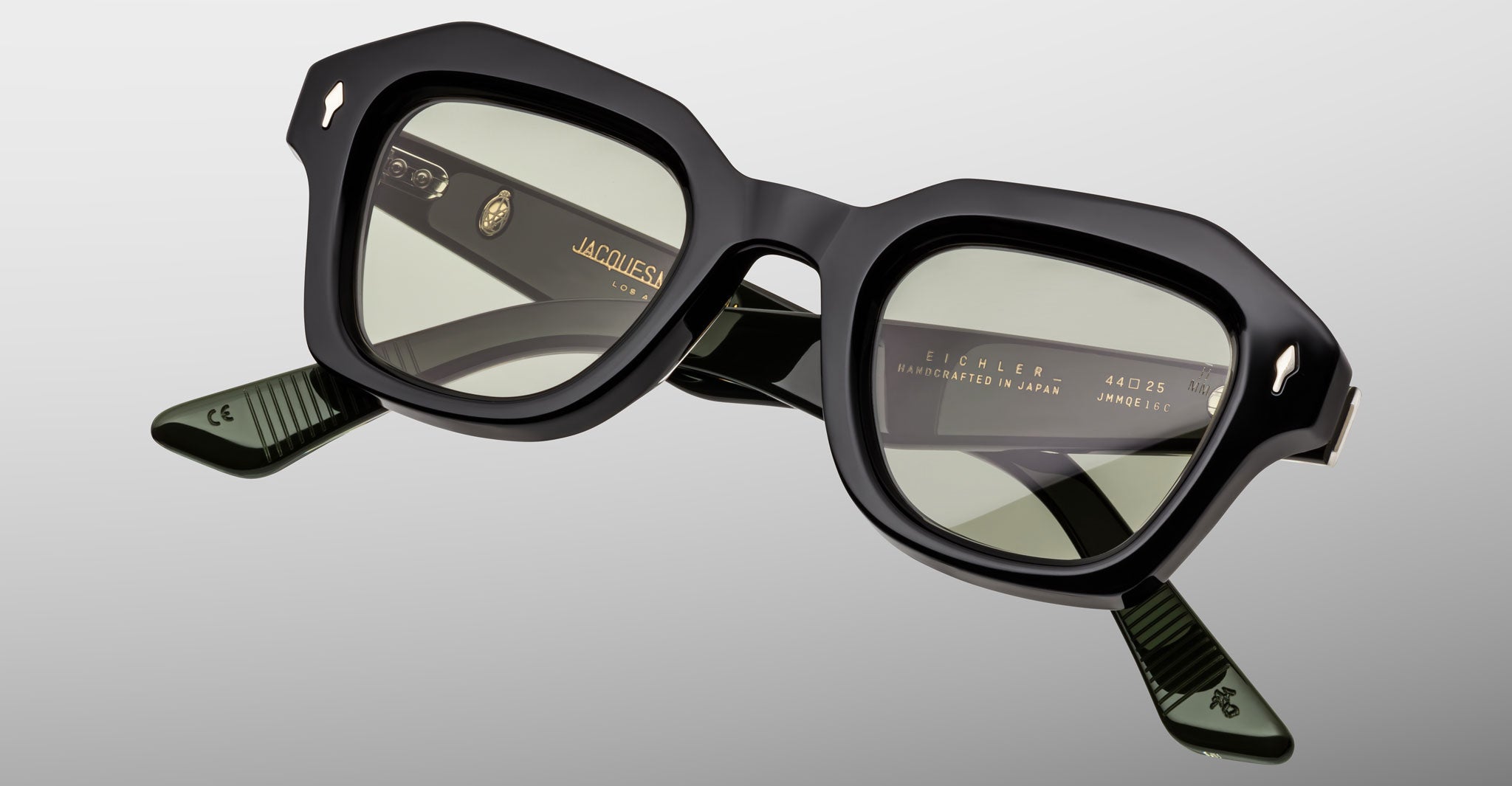 The Jacques Marie Mage Eichler features thick, geometric black frames with green-tinted lenses, shown on a light gray background. Gold lettering highlights the inside of the arms and bridge.