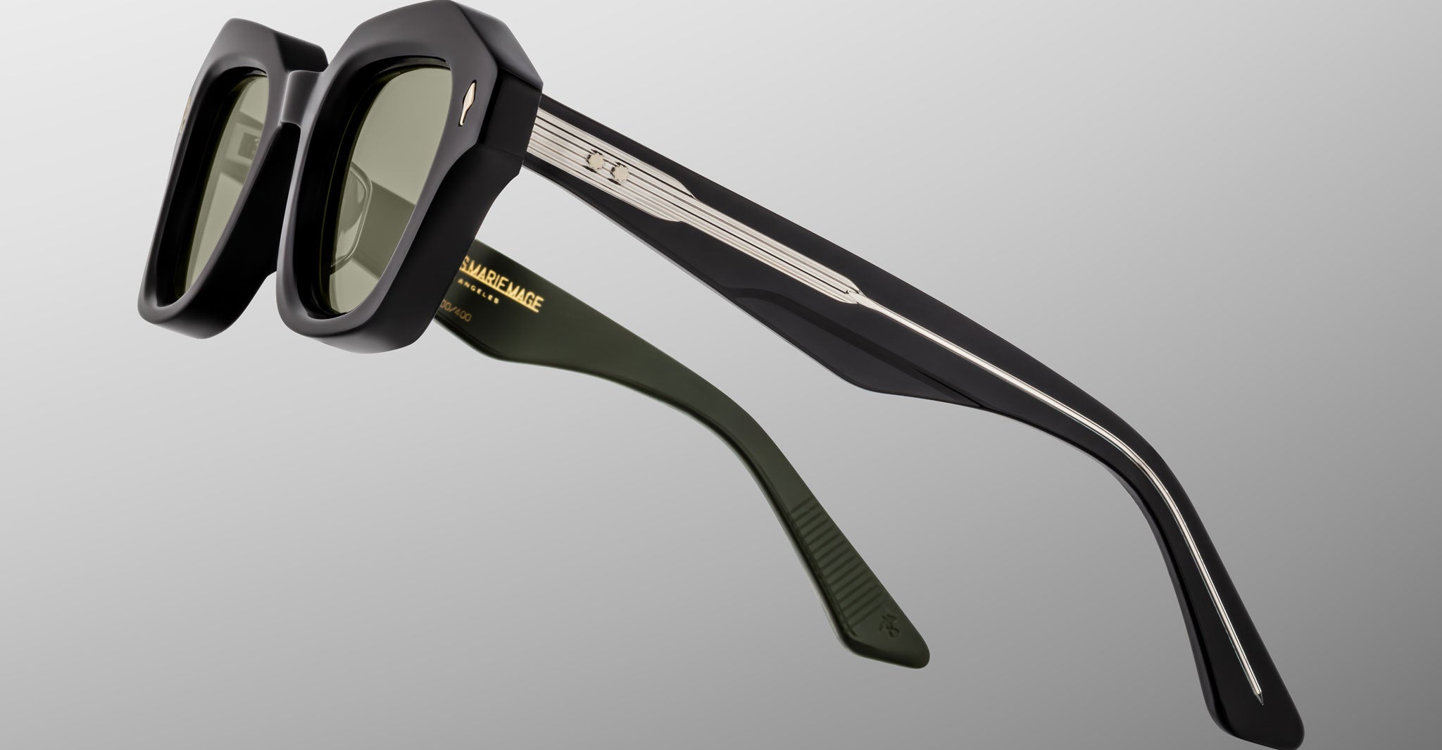 A close-up of Jacques Marie Mage Eichler sunglasses featuring black square frames, green-tinted lenses, and thick temples—one in dark green with gold text—set against a plain light gray background.