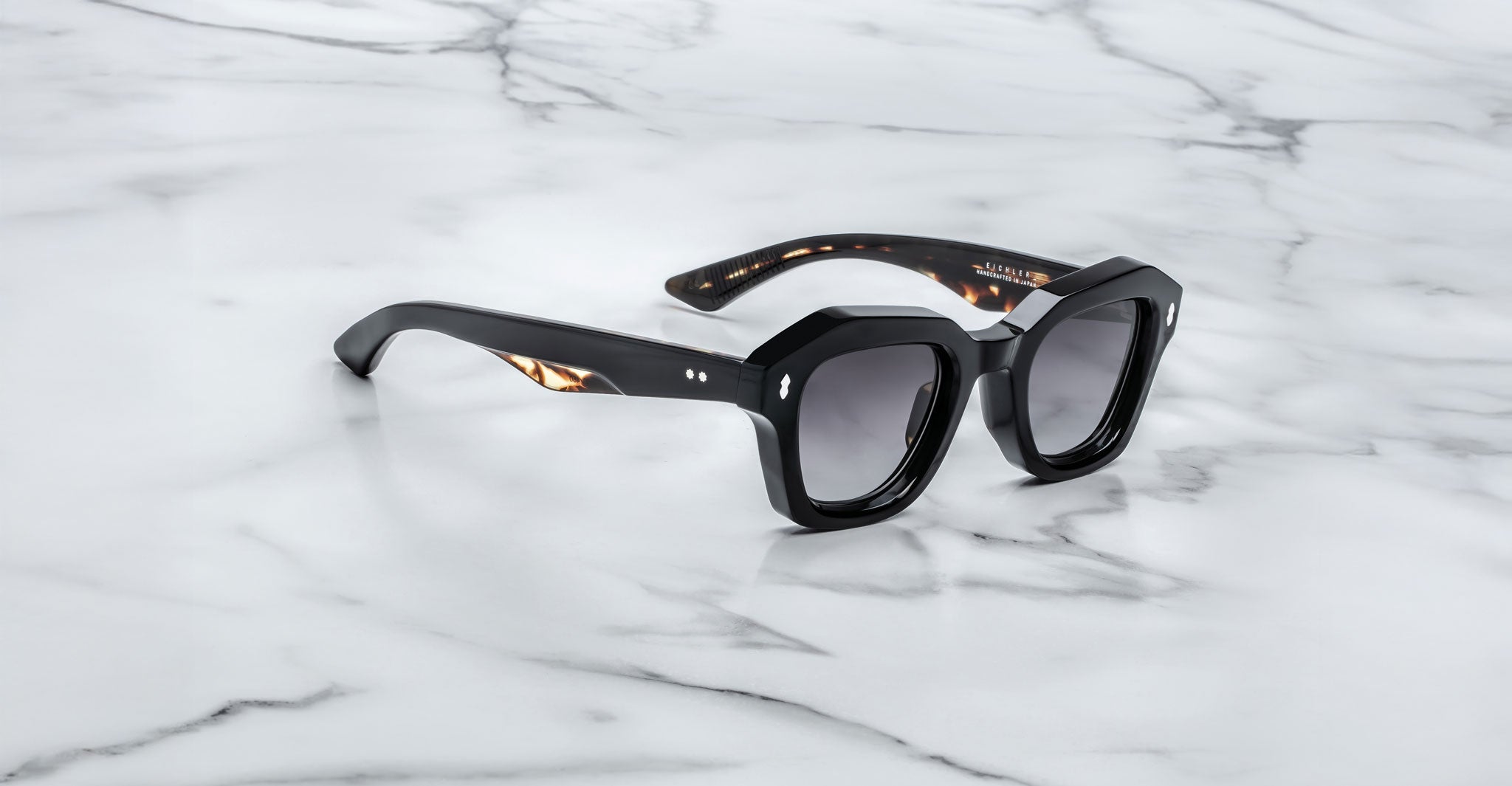 A pair of Jacques Marie Mage Eichler black, thick-framed sunglasses with grey lenses sits on a white marble surface with grey veins