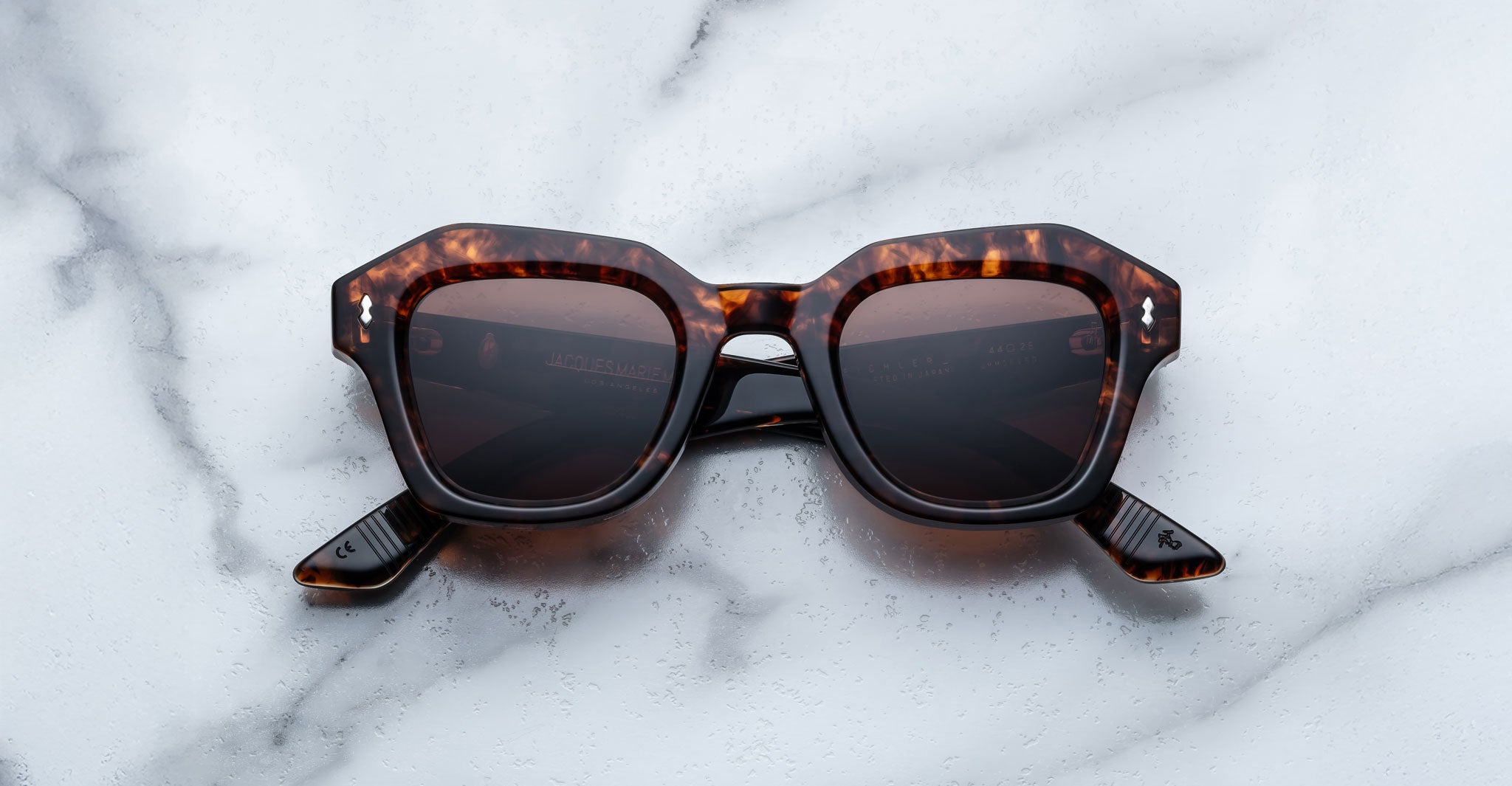 A pair of Jacques Marie Mage Eichler Havana color, thick-framed sunglasses with brown lenses rests on a white marble surface with grey veins, with faint brand details visible on the inside of the left lens.