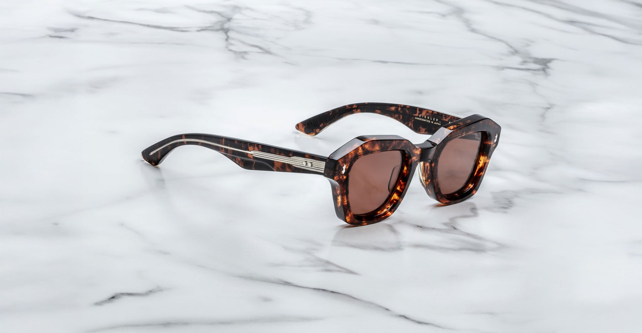 A pair of Jacques Marie Mage Eichler Havana, thick-framed sunglasses with brown lenses sits on a white marble surface with grey veins
