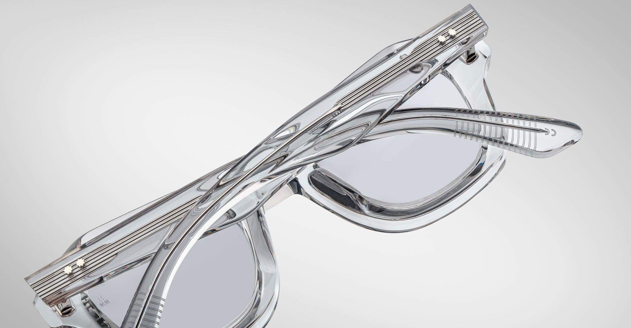 A pair of Jacques Marie Mage Eichler crystal, thick-framed sunglasses with light grey lenses rests on a white marble surface with grey veins, with faint brand details visible on the inside of the left lens.