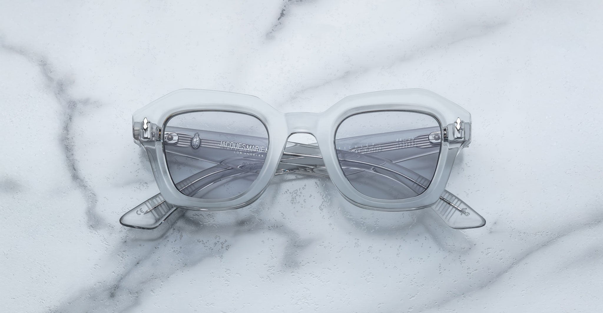 A pair of Jacques Marie Mage Eichler crystal, thick-framed sunglasses with grey lenses rests on a white marble surface with light grey veins, with faint brand details visible on the inside of the left lens.