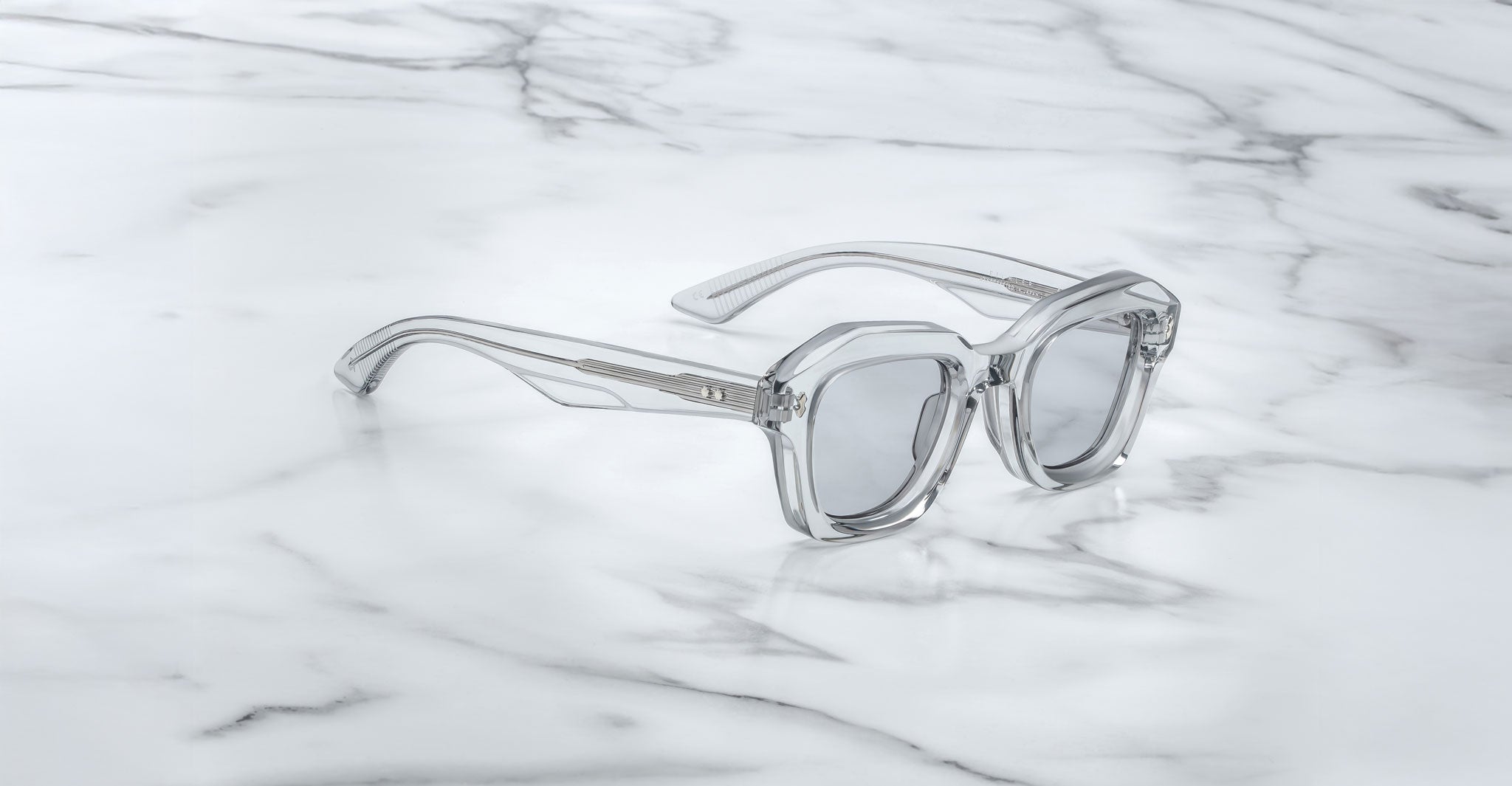 Jacques Marie Mage Eichler sunglasses, featuring clear geometric frames and silver-tinted lenses, rest on a white marble surface with gray veining.