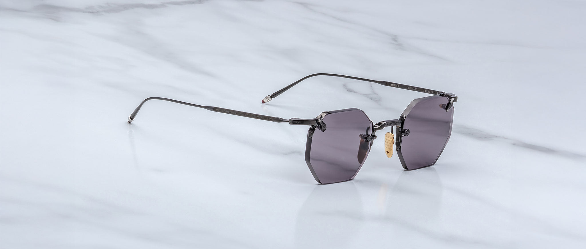 The Jacques Marie Mage El Dorado features geometric rimless sunglasses with dark tinted lenses and slim metal arms, displayed on a light gray marble surface.
