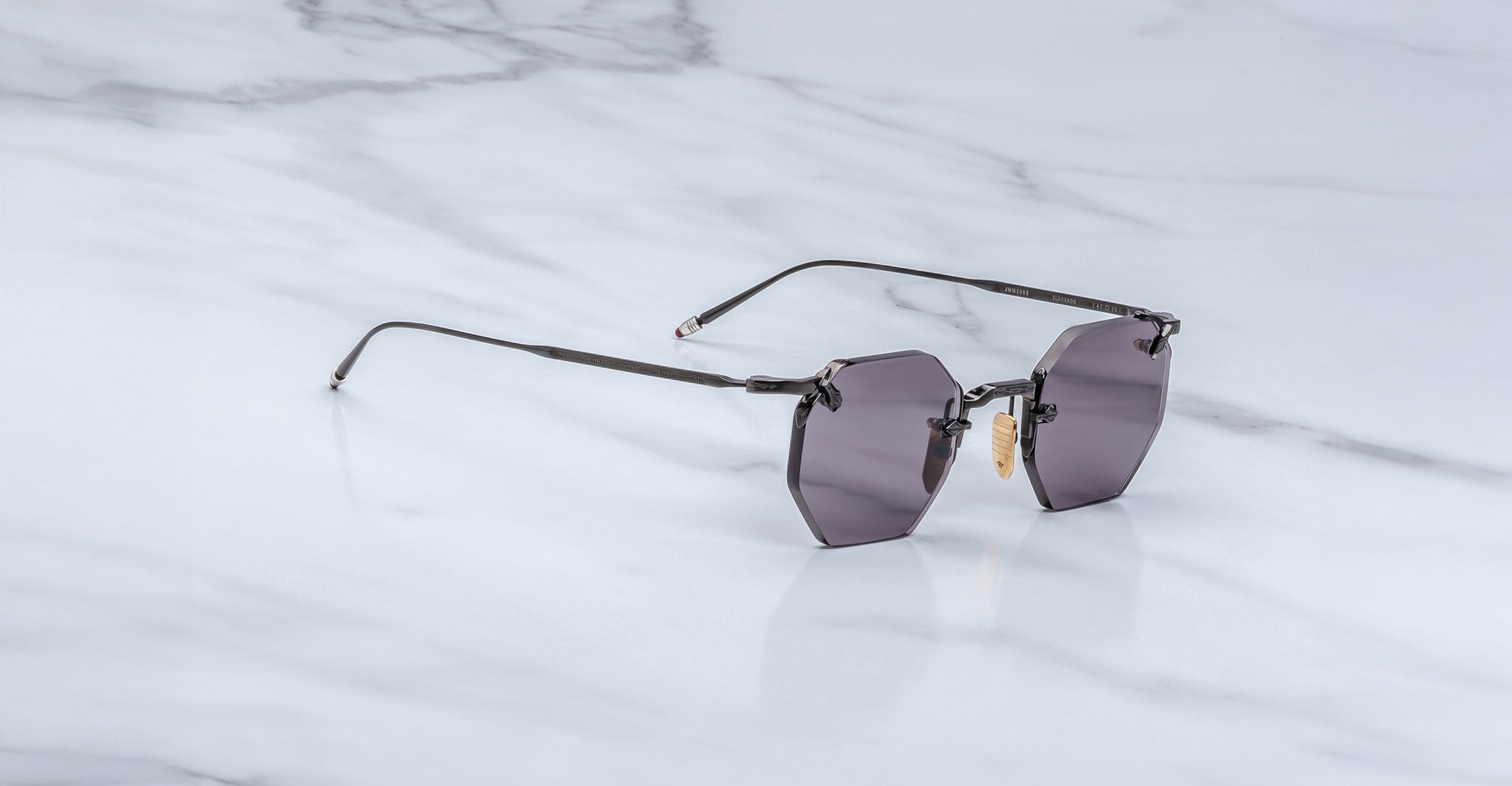 The Jacques Marie Mage El Dorado features geometric rimless sunglasses with dark tinted lenses and slim metal arms, displayed on a light gray marble surface.