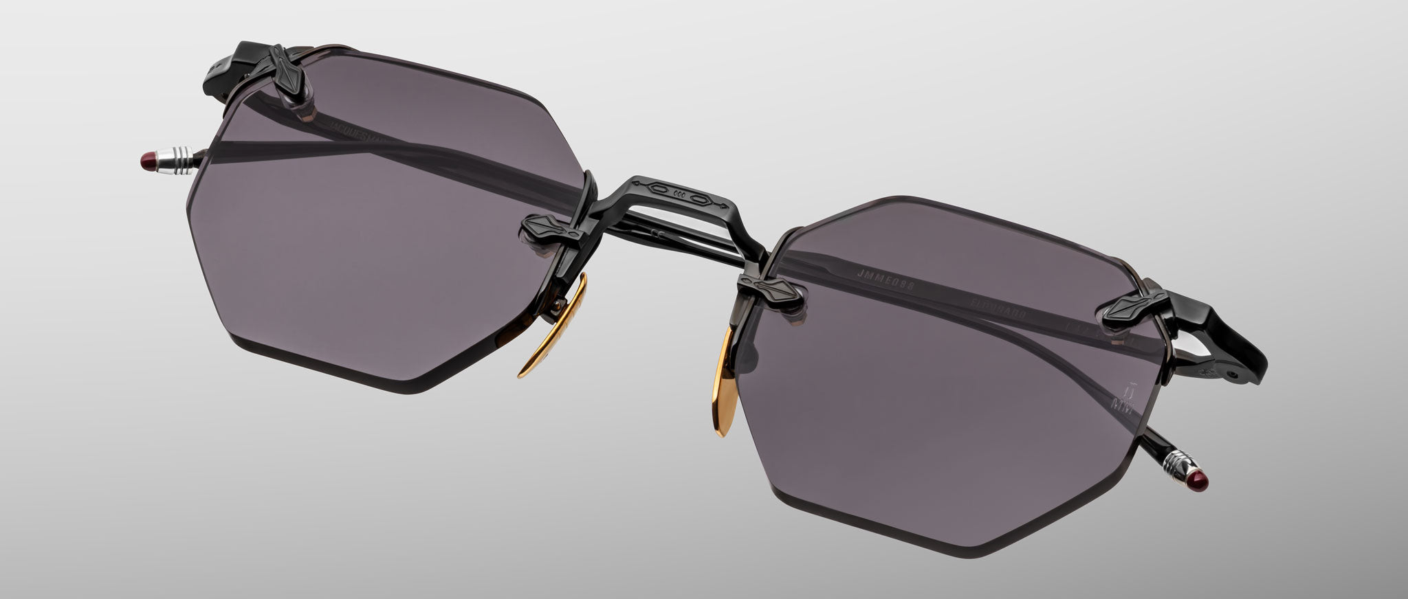 The Jacques Marie Mage El Dorado sunglasses feature geometric hexagonal dark-tinted lenses, thin black metal frames, and gold nose pad accents, showcased on a light gray background.